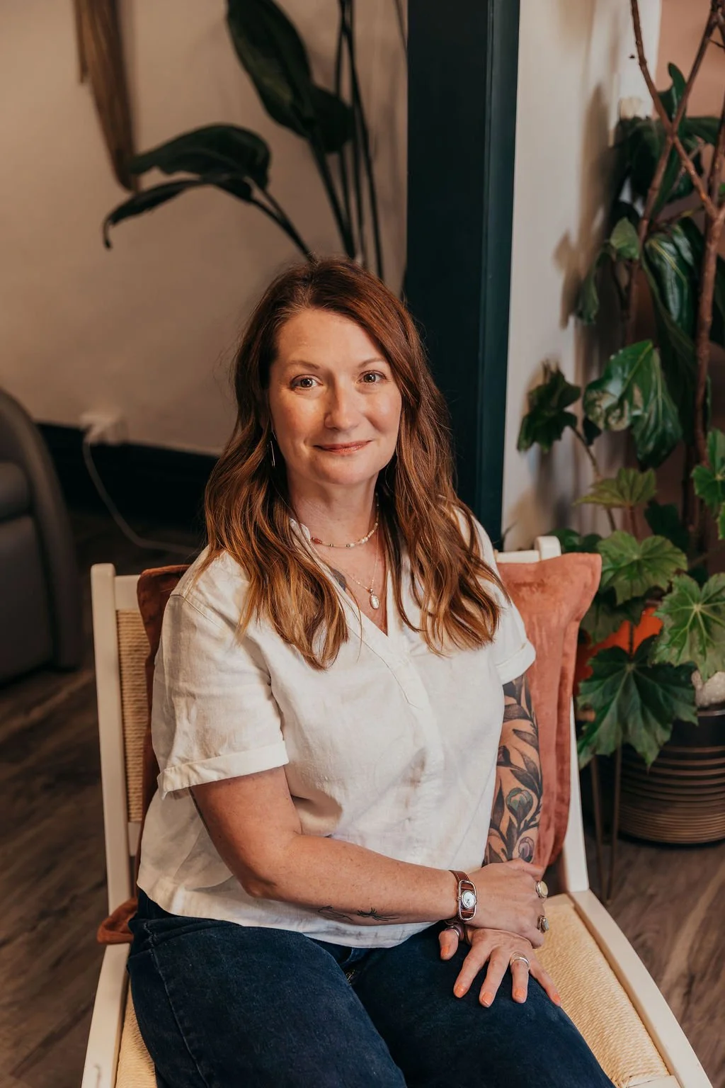 A woman with brown hair wearing a cream-colored blouse and jeans, sitting on a chair with a beige cushion, surrounded by green plants.
