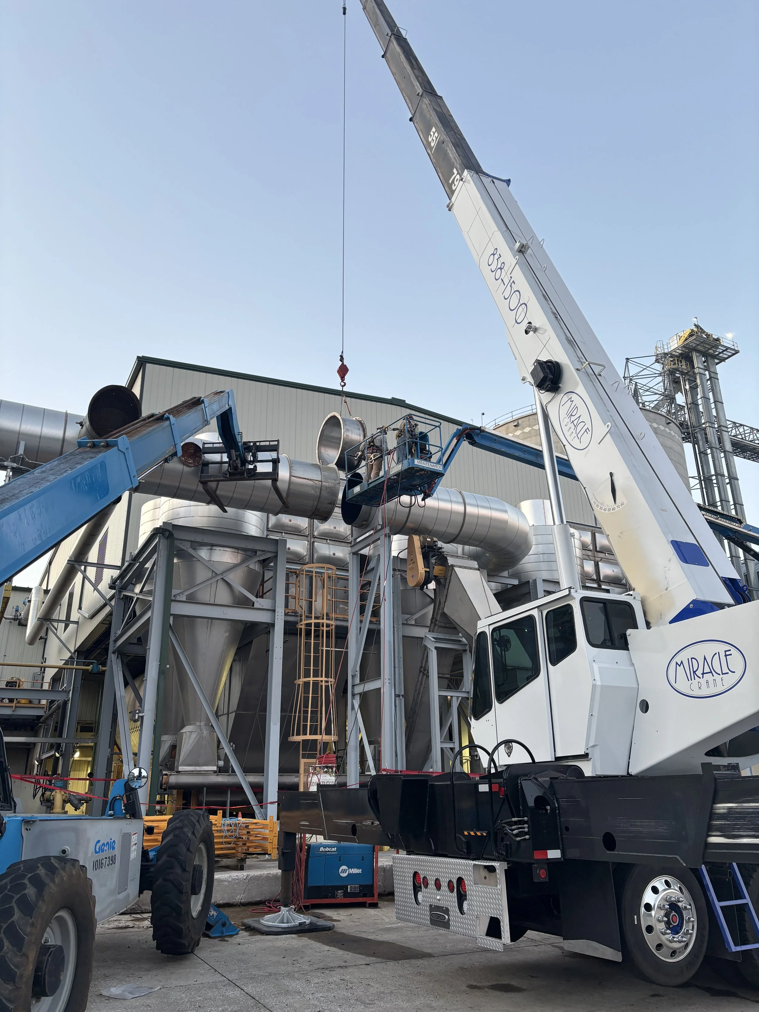 Miracle Crane mobile crane assisting with industrial ductwork lift and installation at manufacturing facility.