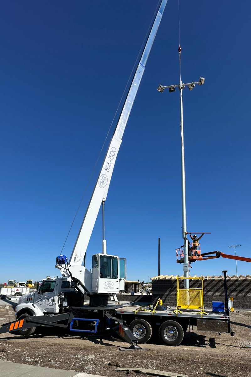 Miracle Crane’s 30-ton crane holding a stadium light pole in position while a worker in a boom lift guides the lower section at Rose Hill High School.