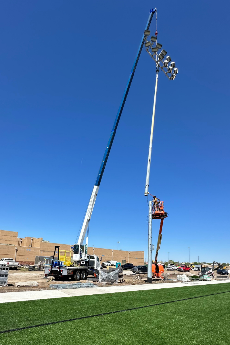 Miracle Crane lifting a stadium light assembly while a worker in a boom lift guides the pole section into place at Rose Hill High School.
