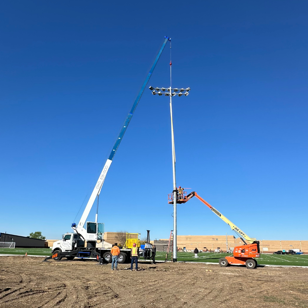 Wide view of Miracle Crane setting a stadium light assembly while a boom lift crew guides the pole at Rose Hill High School.