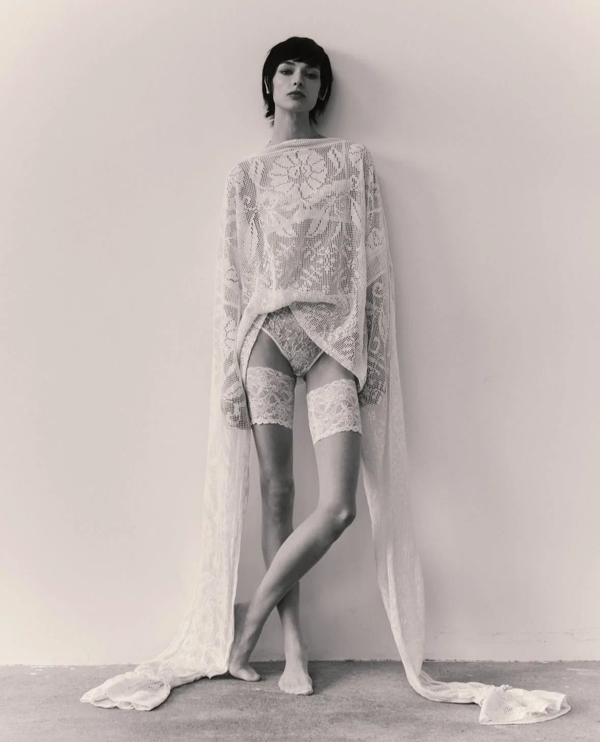 Black and white photo of a woman with short dark hair, wearing a long, lace cardigan over lingerie, standing barefoot against a plain wall.