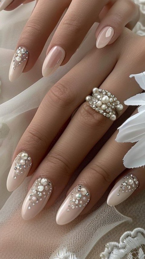 Close-up of hands with elaborately decorated nails and a large pearl and rhinestone ring. The nails are painted in a light, nude color with rhinestone and pearl embellishments.
