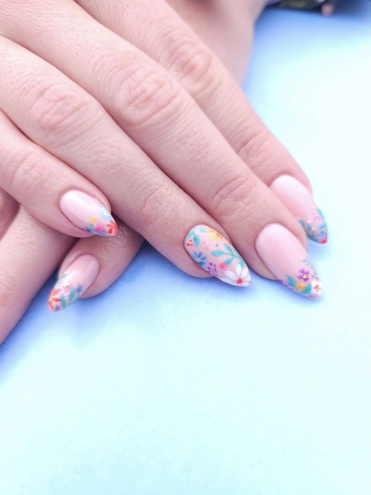 Close-up of manicured fingernails with pastel pink base and colorful floral nail art on a light blue background.