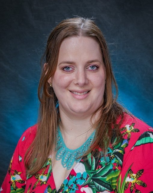 Portrait of a woman with long brown hair, blue eyes, and light skin, smiling, wearing a vibrant pink and green floral top and a turquoise necklace, against a blue gradient background.