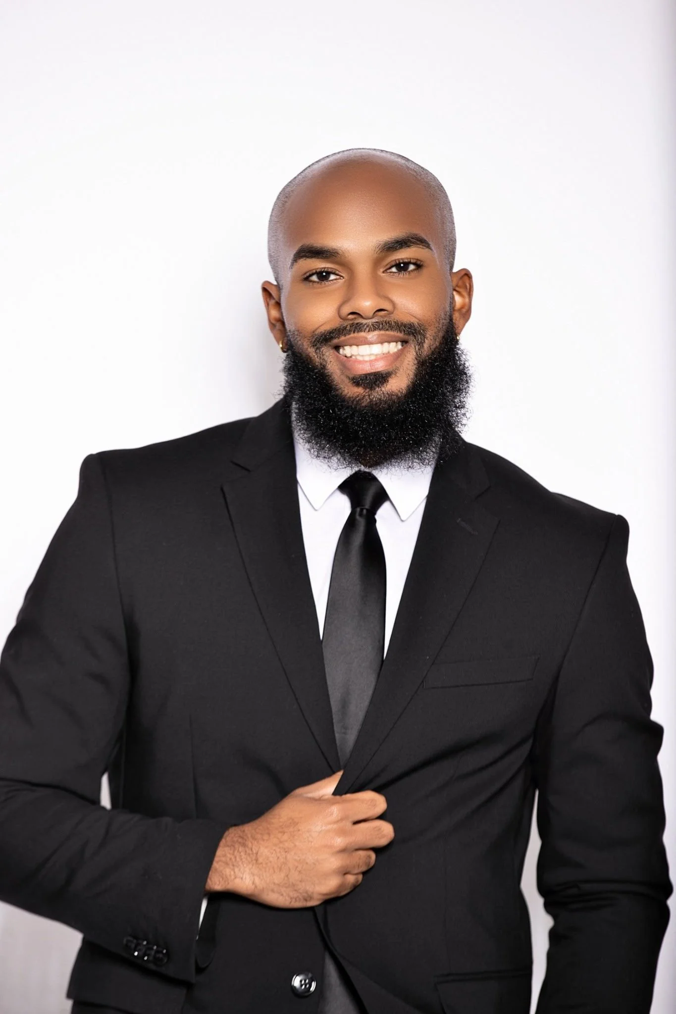 A smiling man with a shaved head and a beard, wearing a black suit, white shirt, and black tie, stands against a plain white background.