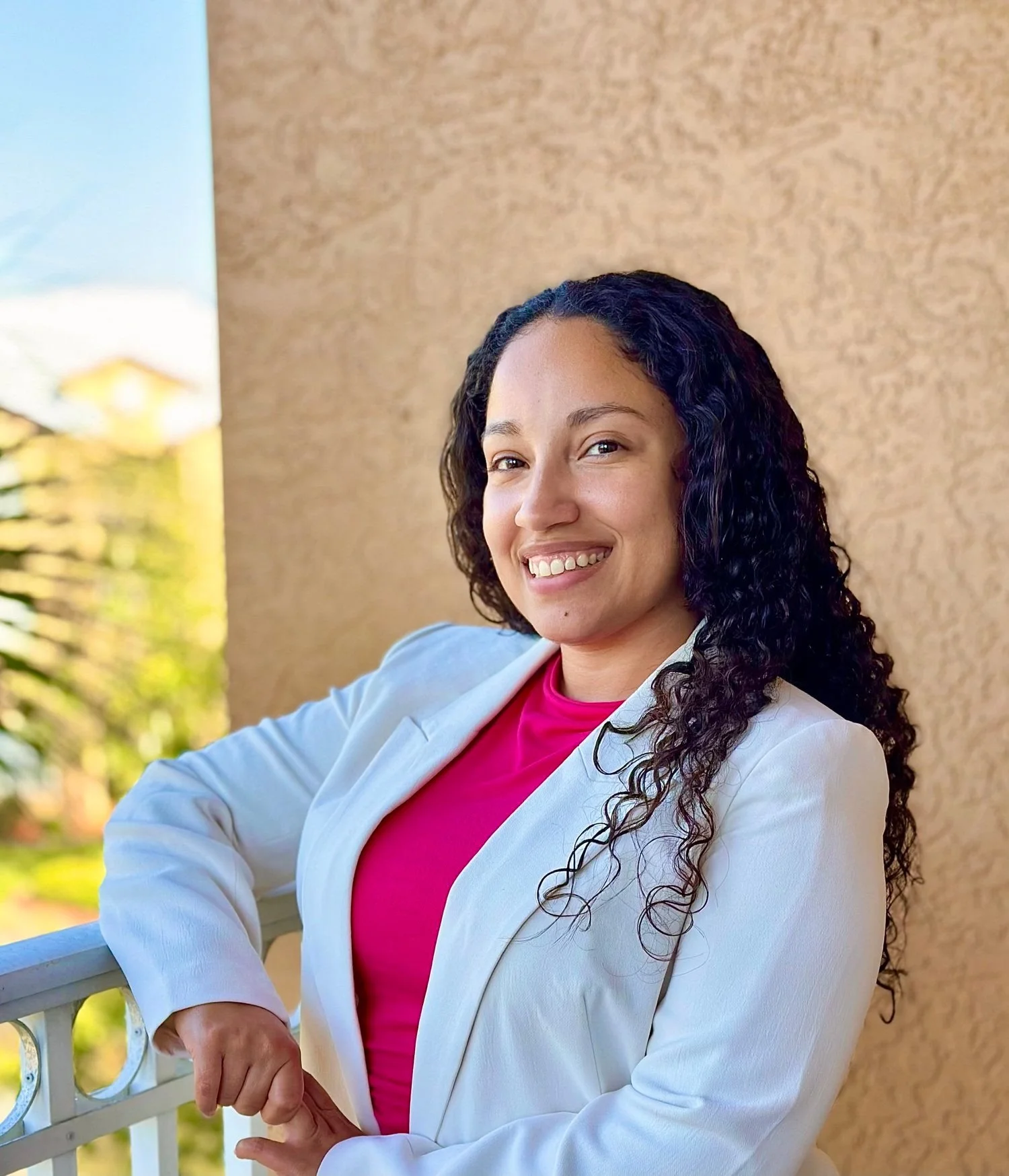 Smiling woman with curly dark hair wearing a white blazer and pink top, standing outdoors near a beige wall and greenery.