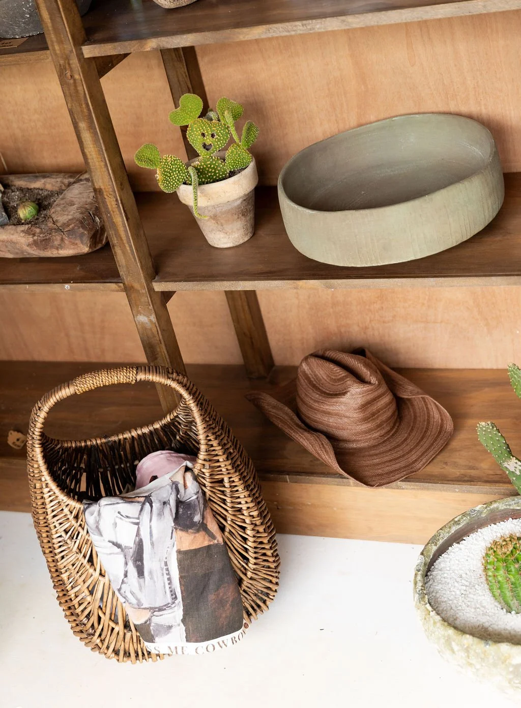 Display shelf with potted cactus, blank ceramic bowl, woven bag with scarf, and brown hat.