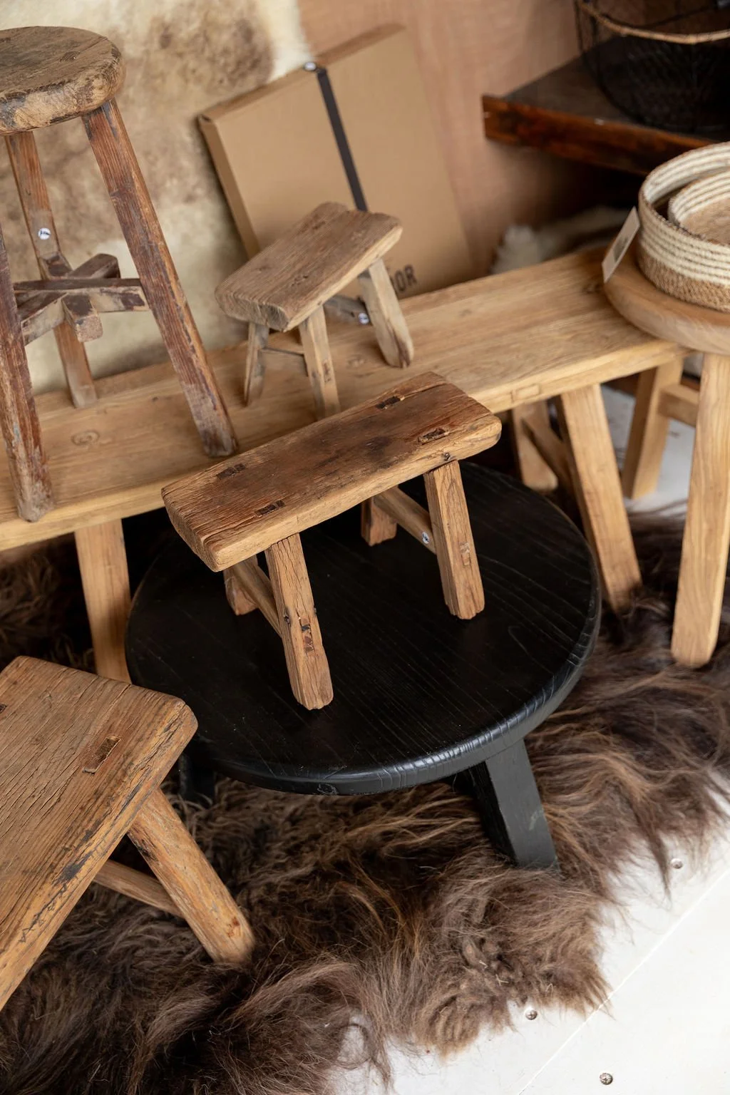 Various small rustic wooden stools and benches displayed on a furry rug, with some stacked or leaning against each other in a store or showroom setting.