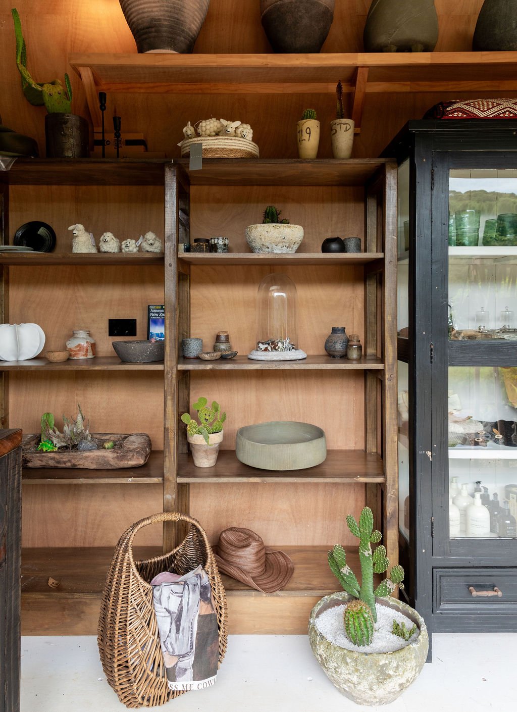 Wooden shelves and display cabinet with decorative items including small cactus plants, pottery, and animal figurines, a wicker bag with clothing inside, and a potted cactus in front of the shelves.