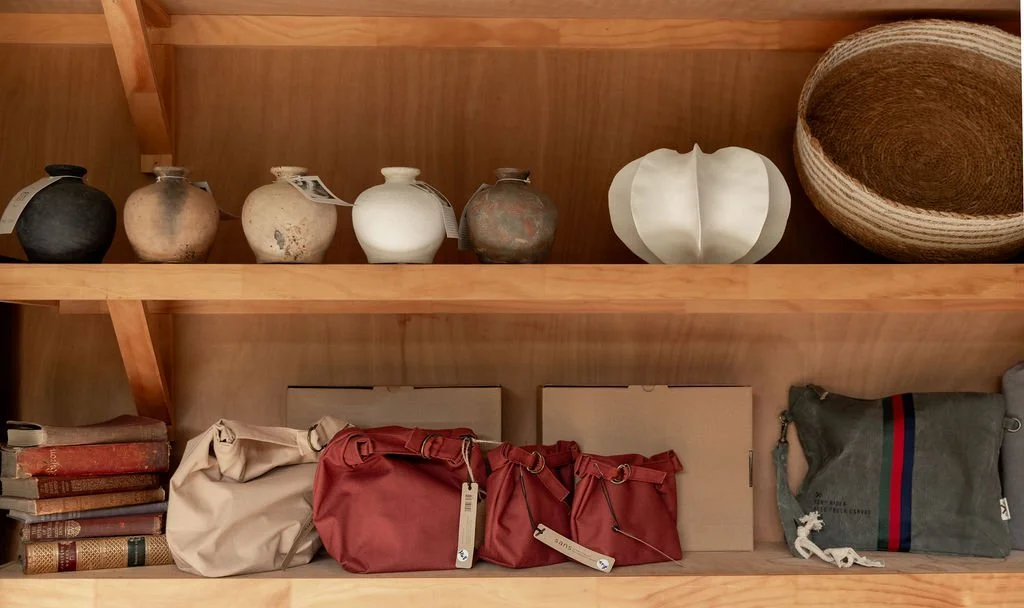 Shelf with collection of ceramic pottery, woven basket, hardcover books, fabric bags, and cardboard boxes.