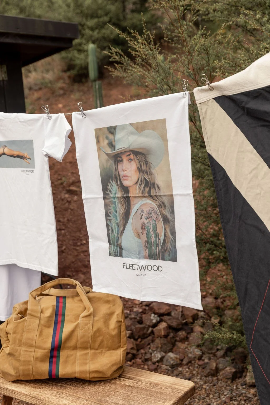 A clothing display featuring Fleetwood band T-shirts hung on a clothesline with clothespins, including a white T-shirt with a woman in a cowboy hat, and a beige bag with a colorful stripe, all set outdoors among desert vegetation.