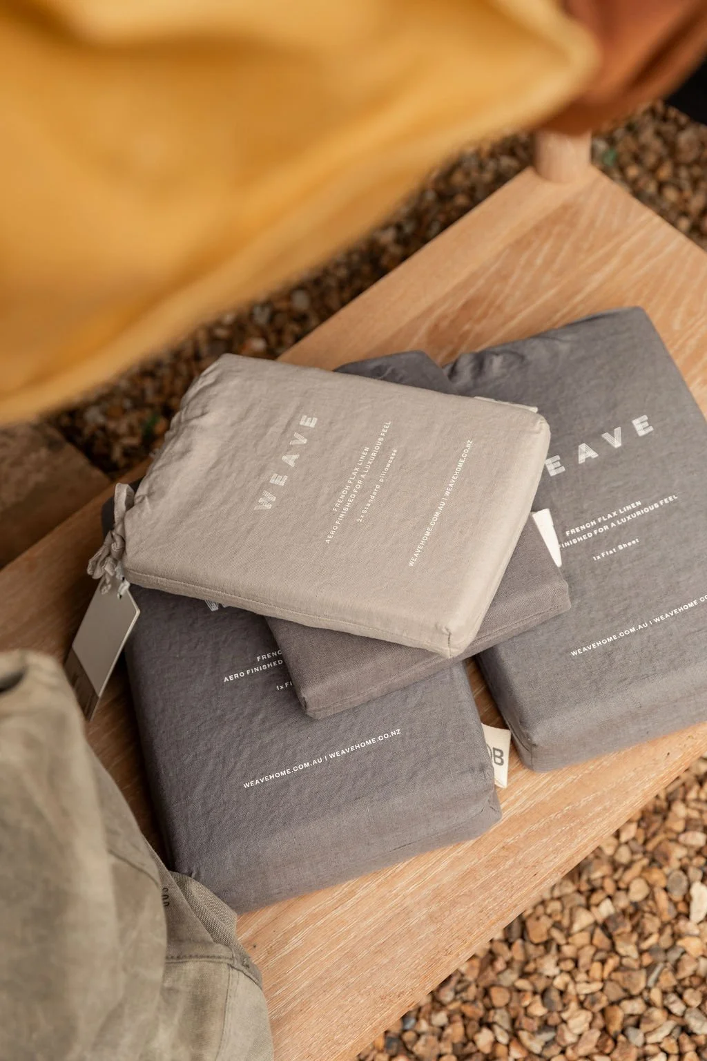 Wrapped and folded textiles on a wooden bench, with gravel underneath, including branded packaging from WEAVEHOME.