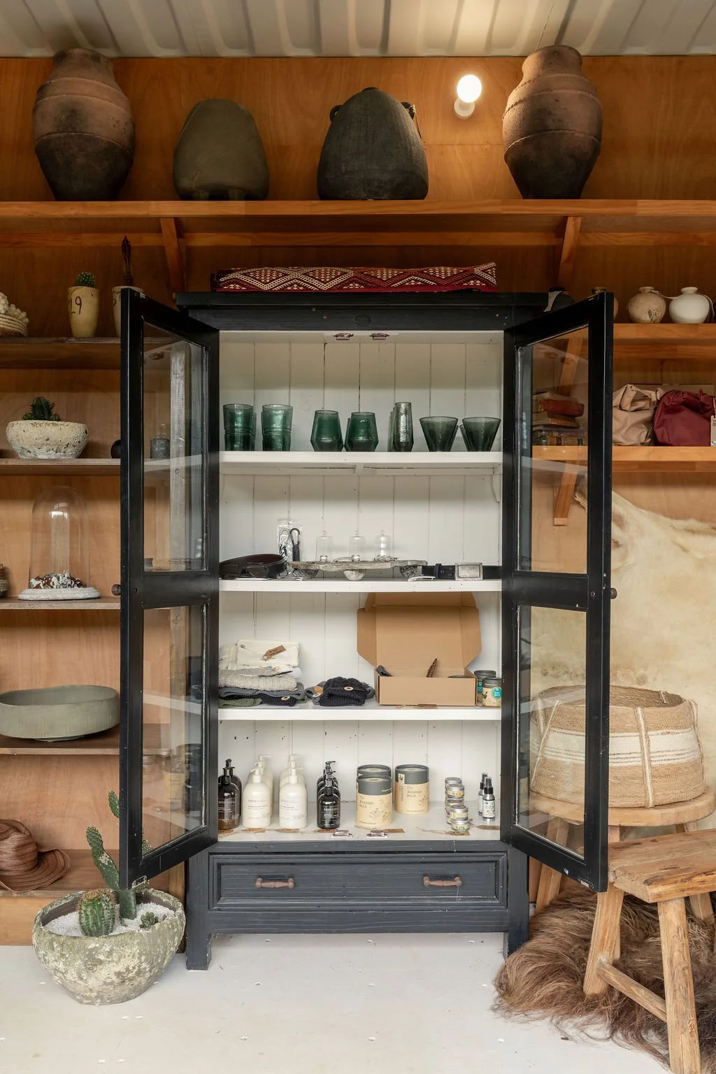 Open black cabinet with glass doors on a white room with wooden walls, displaying green glasses, bottles, and miscellaneous items, with decorative pottery on shelves and a potted cactus in front.