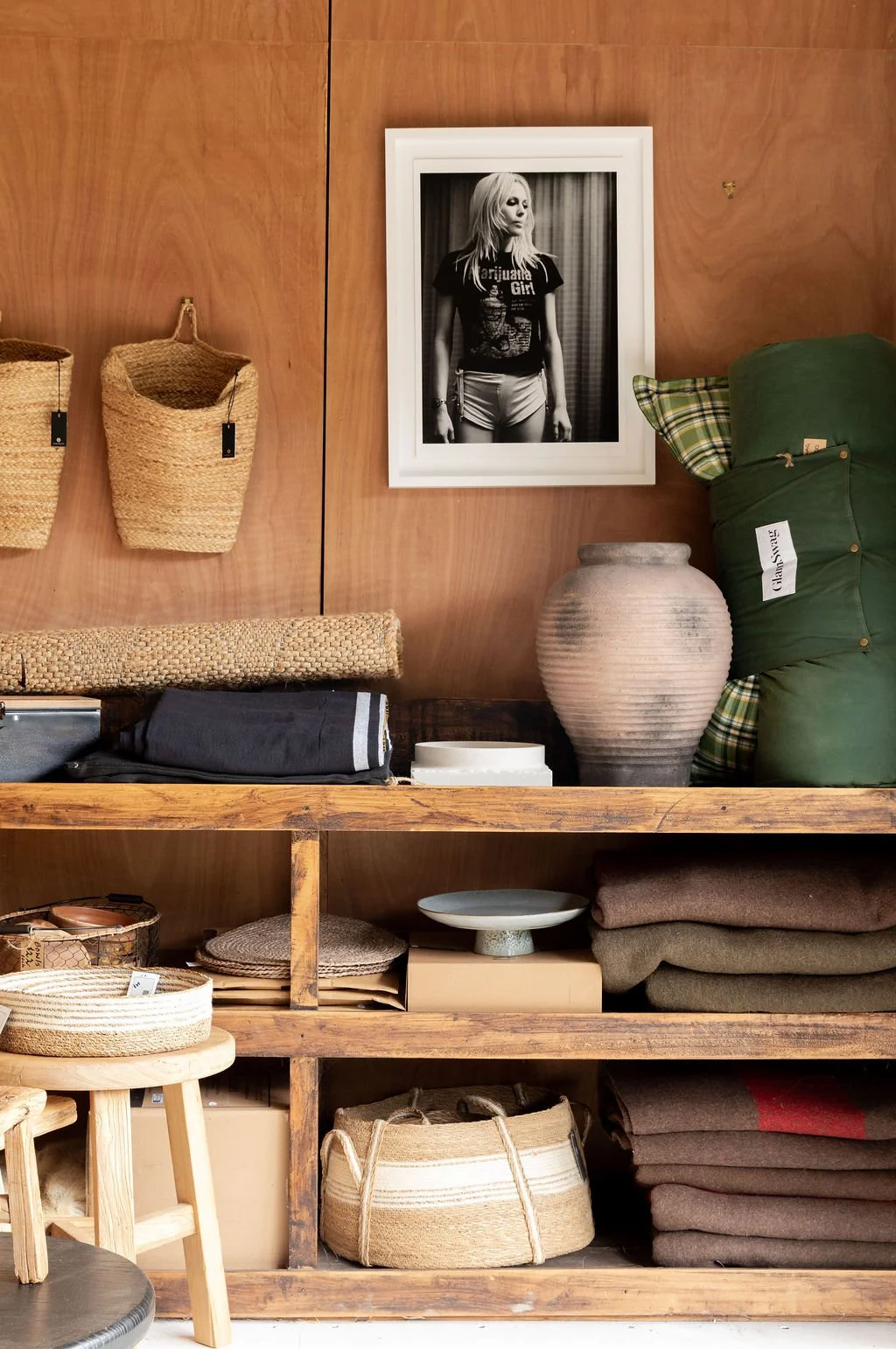 Wooden shelf with woven baskets, folded clothing, ceramic vase, and decorative items, with a black and white framed photo of a woman hanging on a wooden wall above.