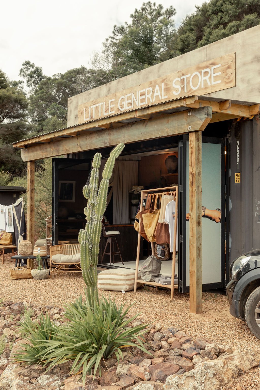 Exterior of a small store called 'Little General Store' with an open front, showing clothing and decor inside, a cactus in the foreground, and trees in the background.
