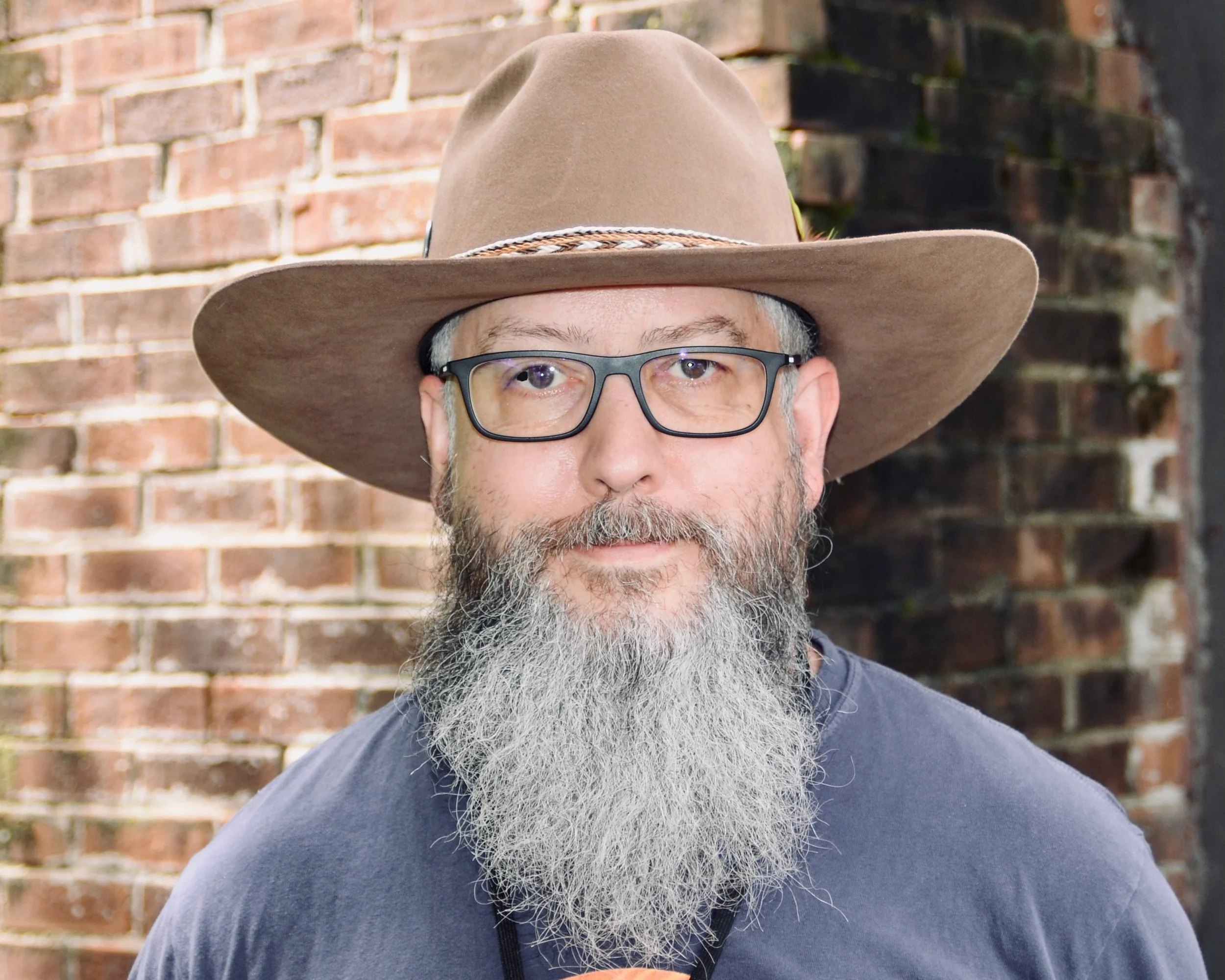 A man with gray hair, a beard, and glasses smiling outdoors with green trees in the background.