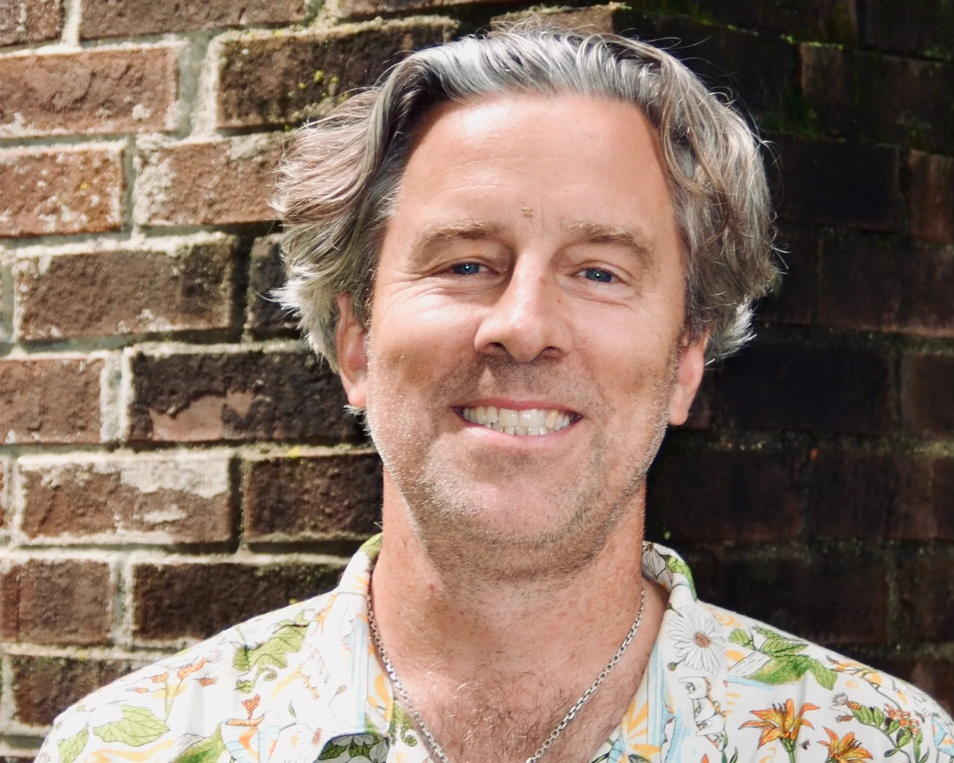 A middle-aged man with gray hair and a beard, smiling, standing in front of a stone wall.