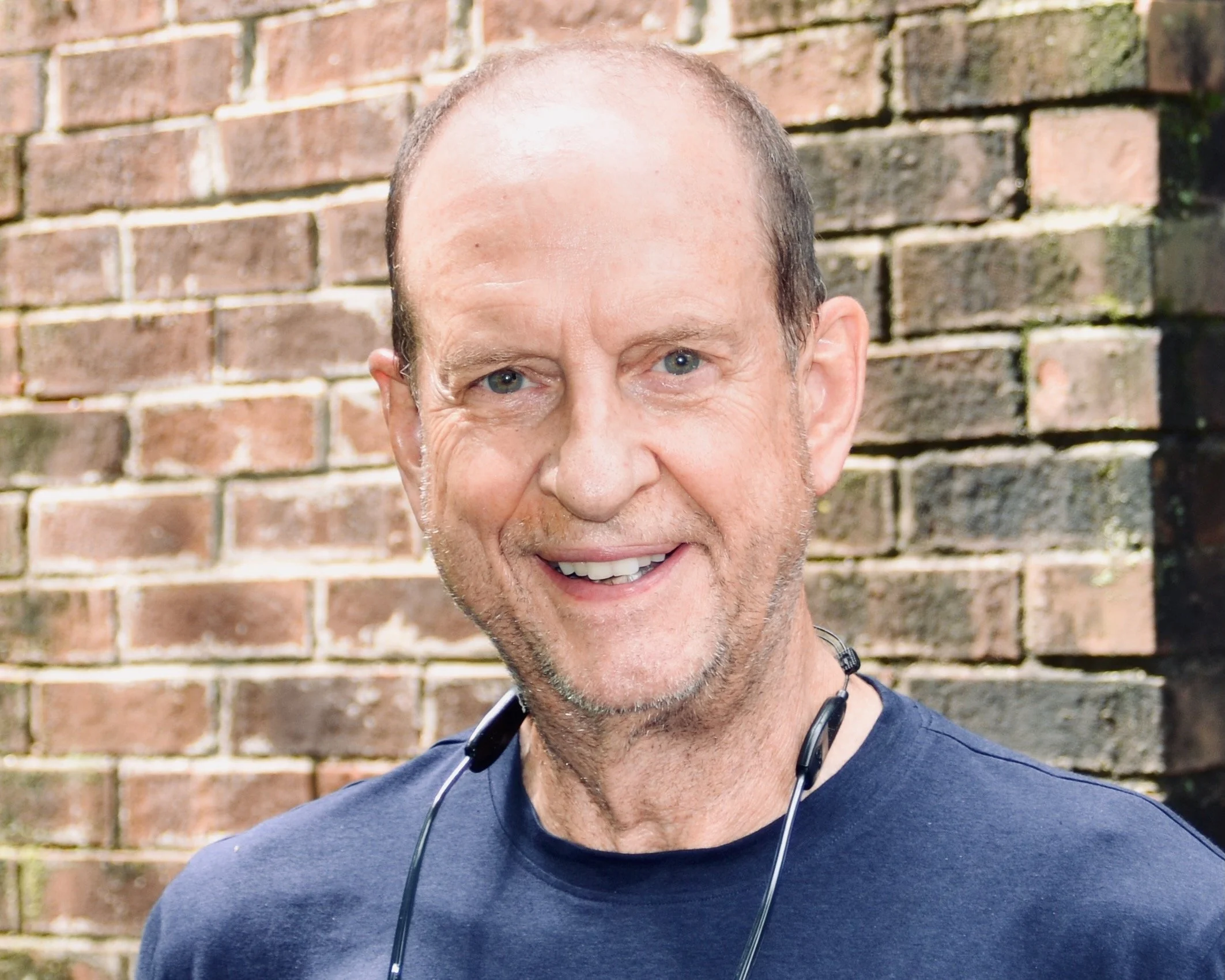 A close-up photo of a middle-aged man with short, receding hair, beard, and expressive eyes, standing outdoors in front of a stone wall.