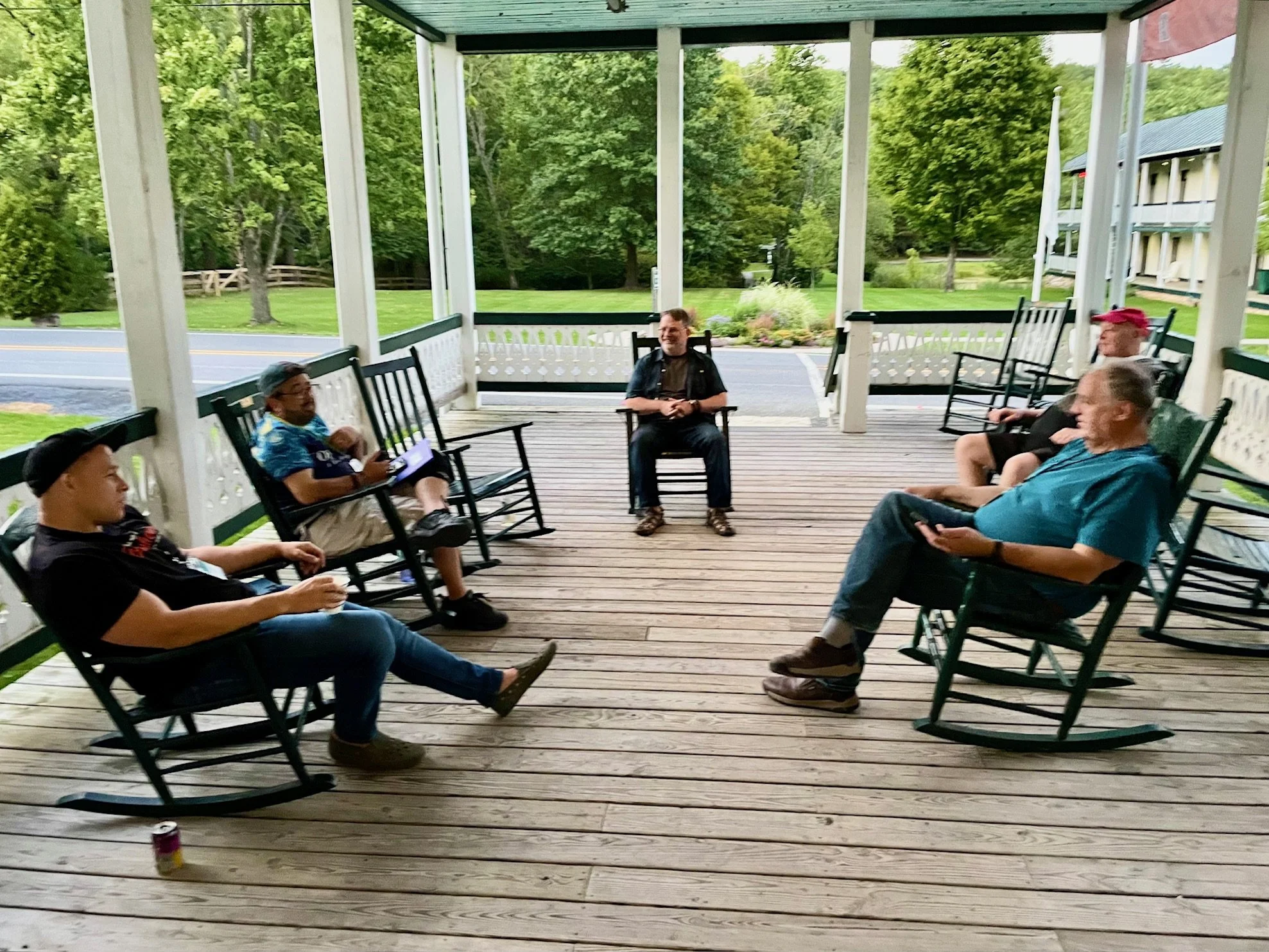 Group of five people sitting on rocking chairs on a wooden porch, surrounded by greenery, having a casual conversation.