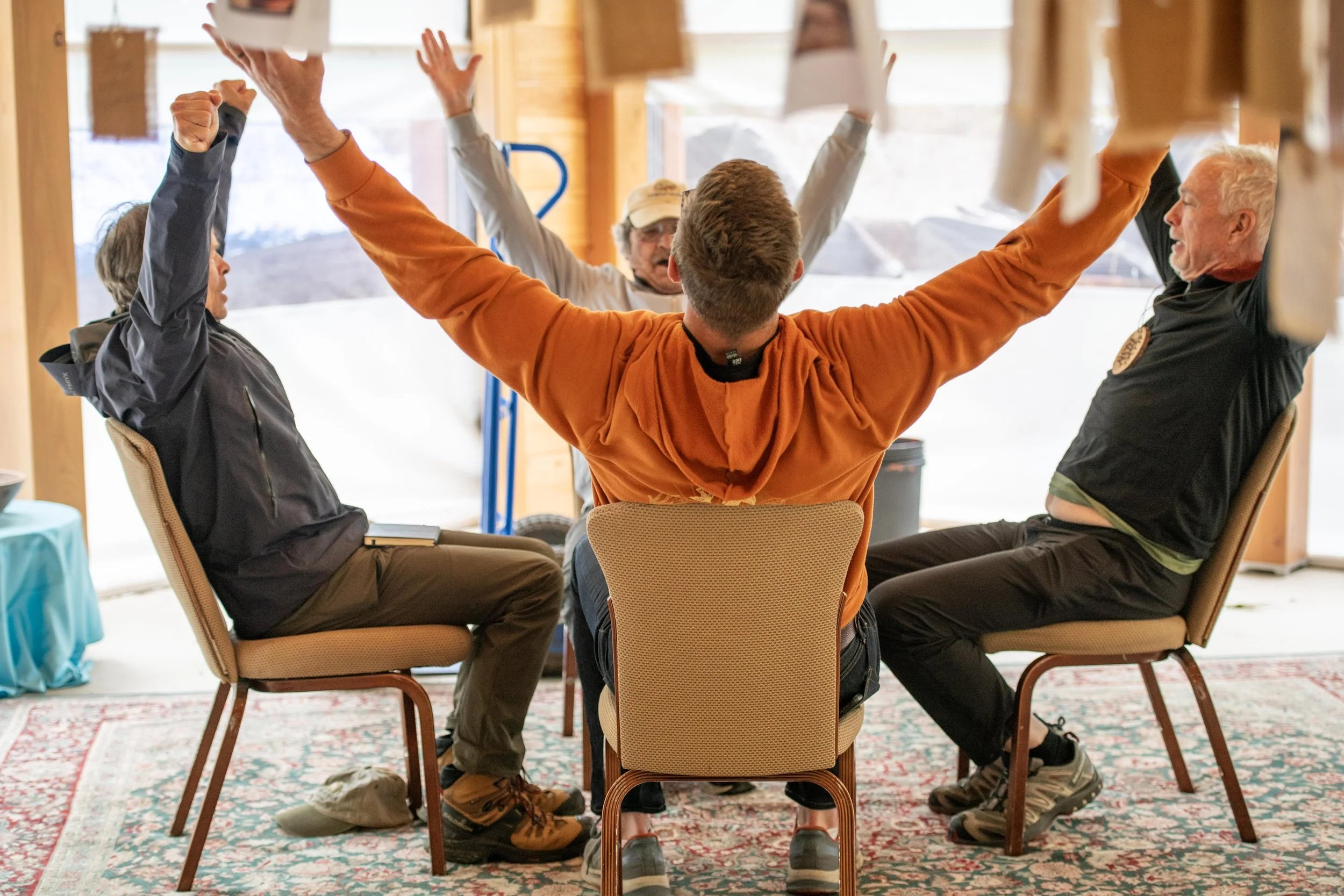 A group of five people sitting in chairs around a table, raising their arms and cheering during a meeting or discussion in a sunlit room with wooden walls and a patterned area rug.