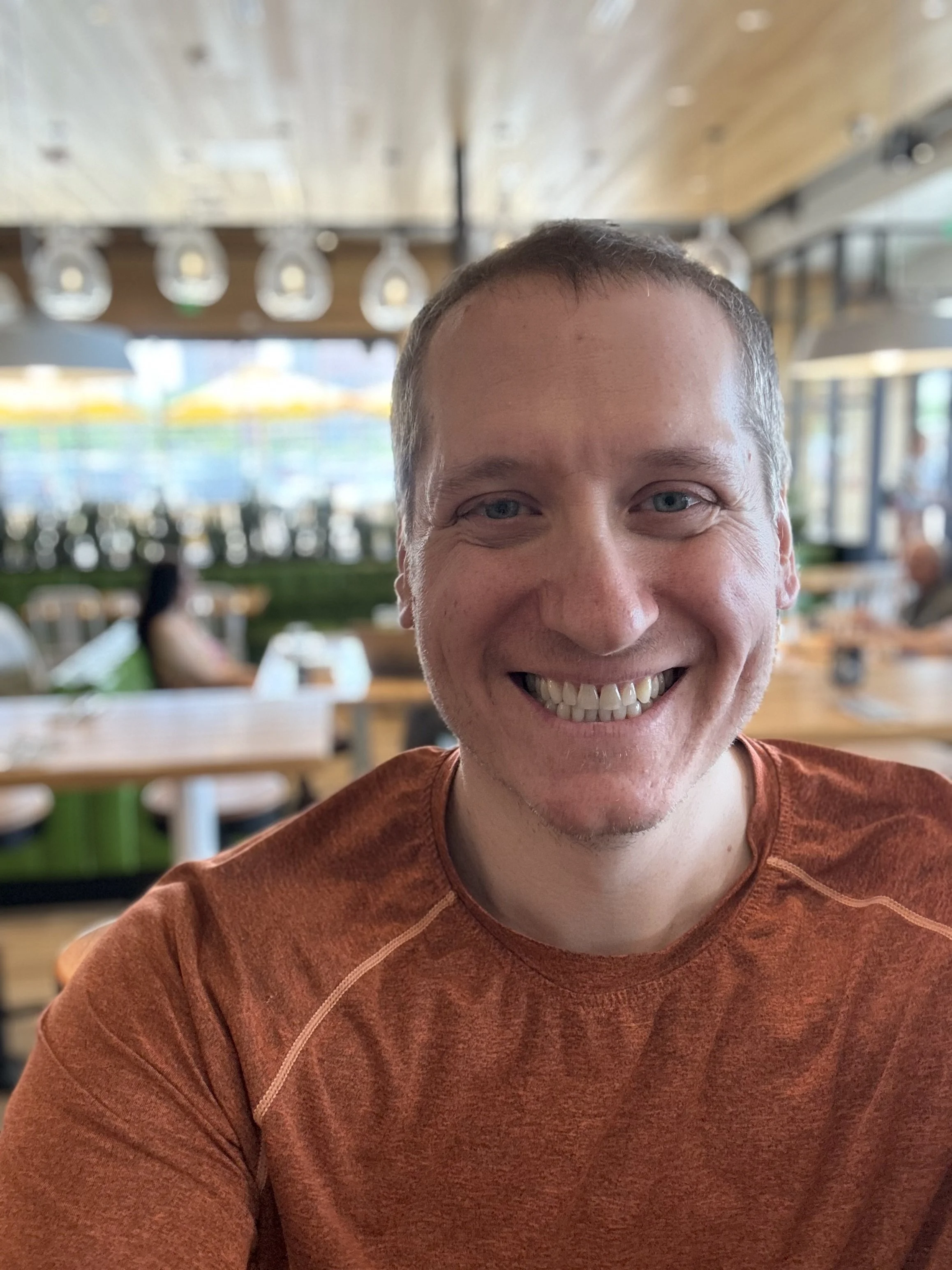 Close-up of a smiling man with short hair wearing a reddish-orange shirt in a restaurant or cafe with wooden ceiling and hanging lights.