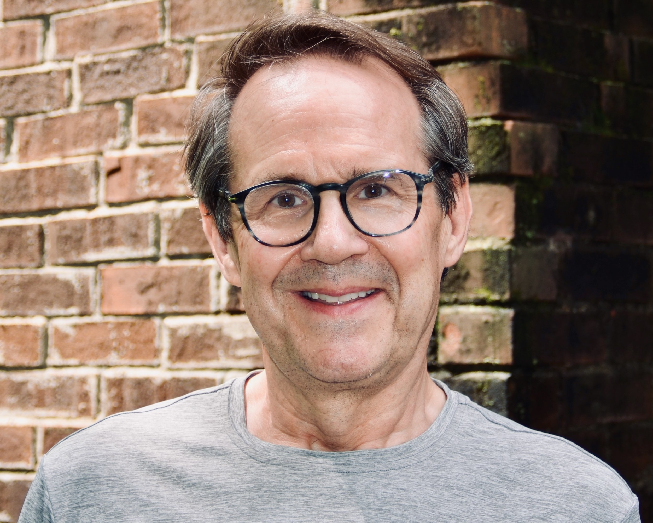 A smiling man wearing glasses and a red jacket standing in front of a stone wall.