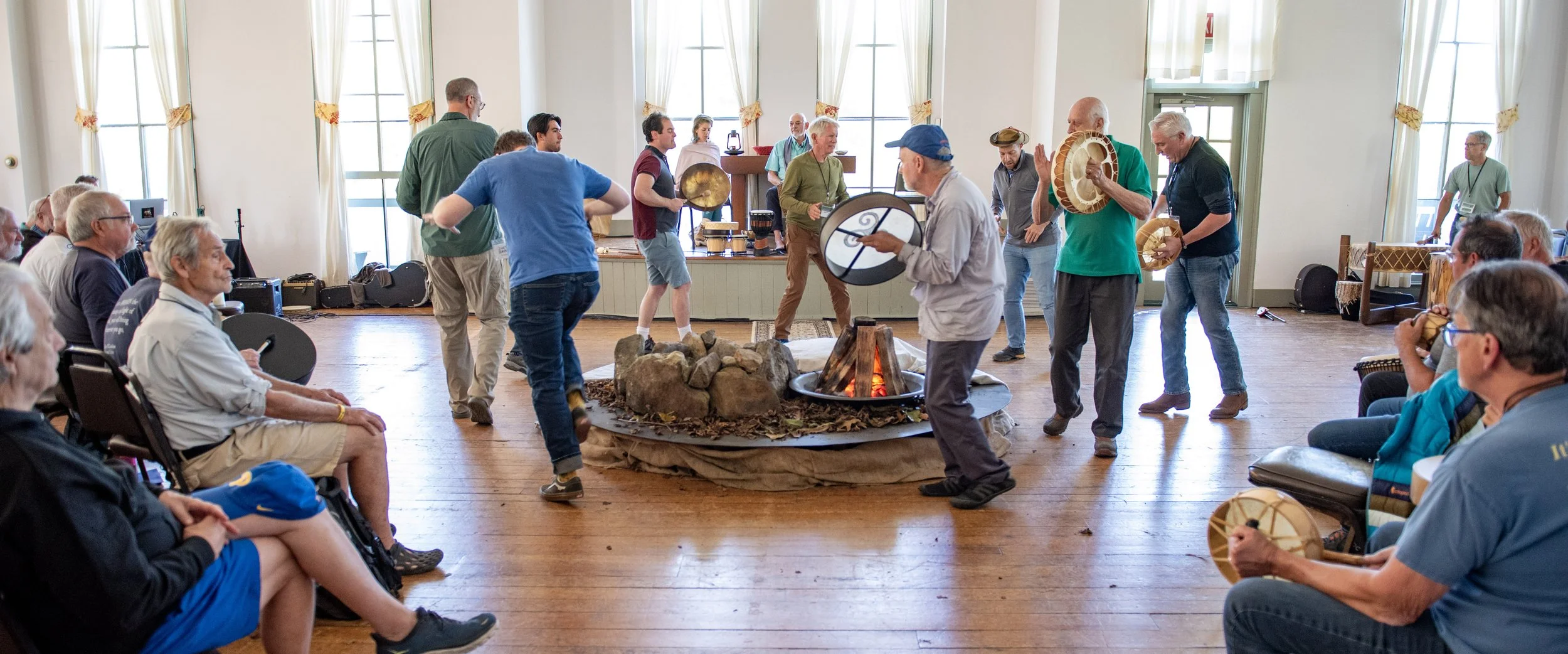 Group of people participating in a drumming circle indoors. Several individuals in the center are holding and playing frame drums around a faux campfire setup, while others watch from seated positions.