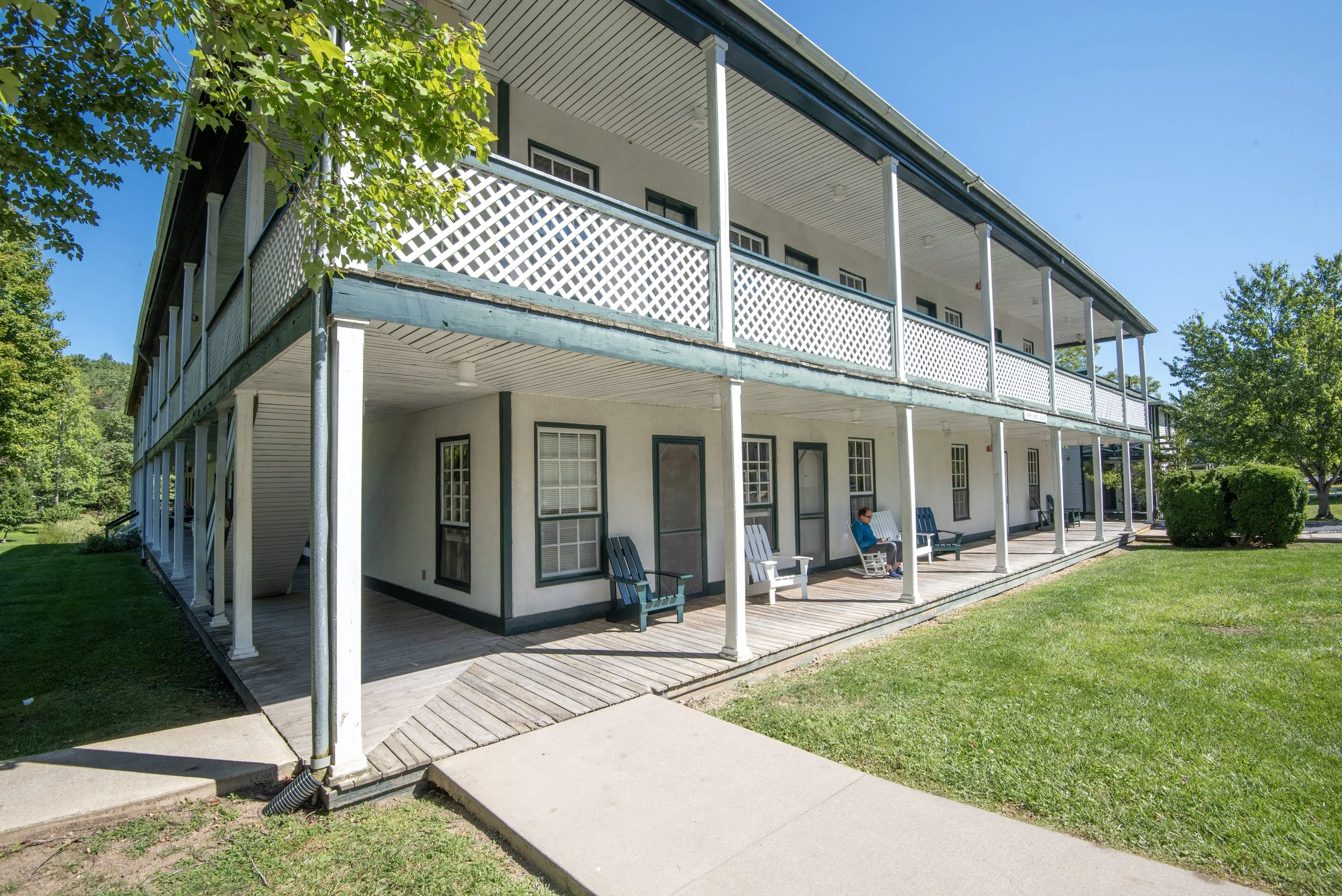 Two-story white building with a porch and rocking chairs, surrounded by green grass and trees, under a clear blue sky.