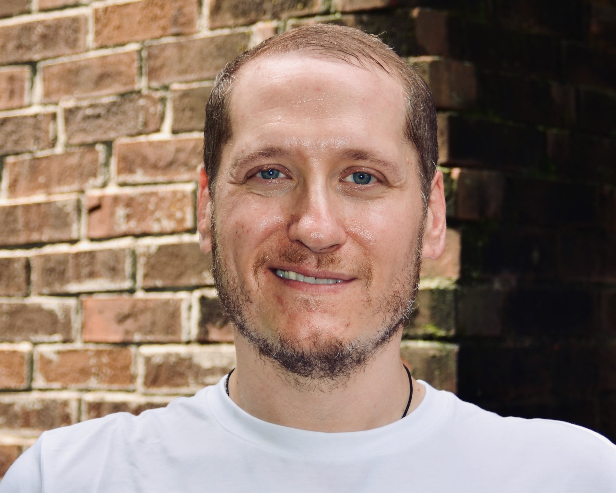 Close-up of a smiling man with short hair wearing a reddish-orange shirt in a restaurant or cafe with wooden ceiling and hanging lights.