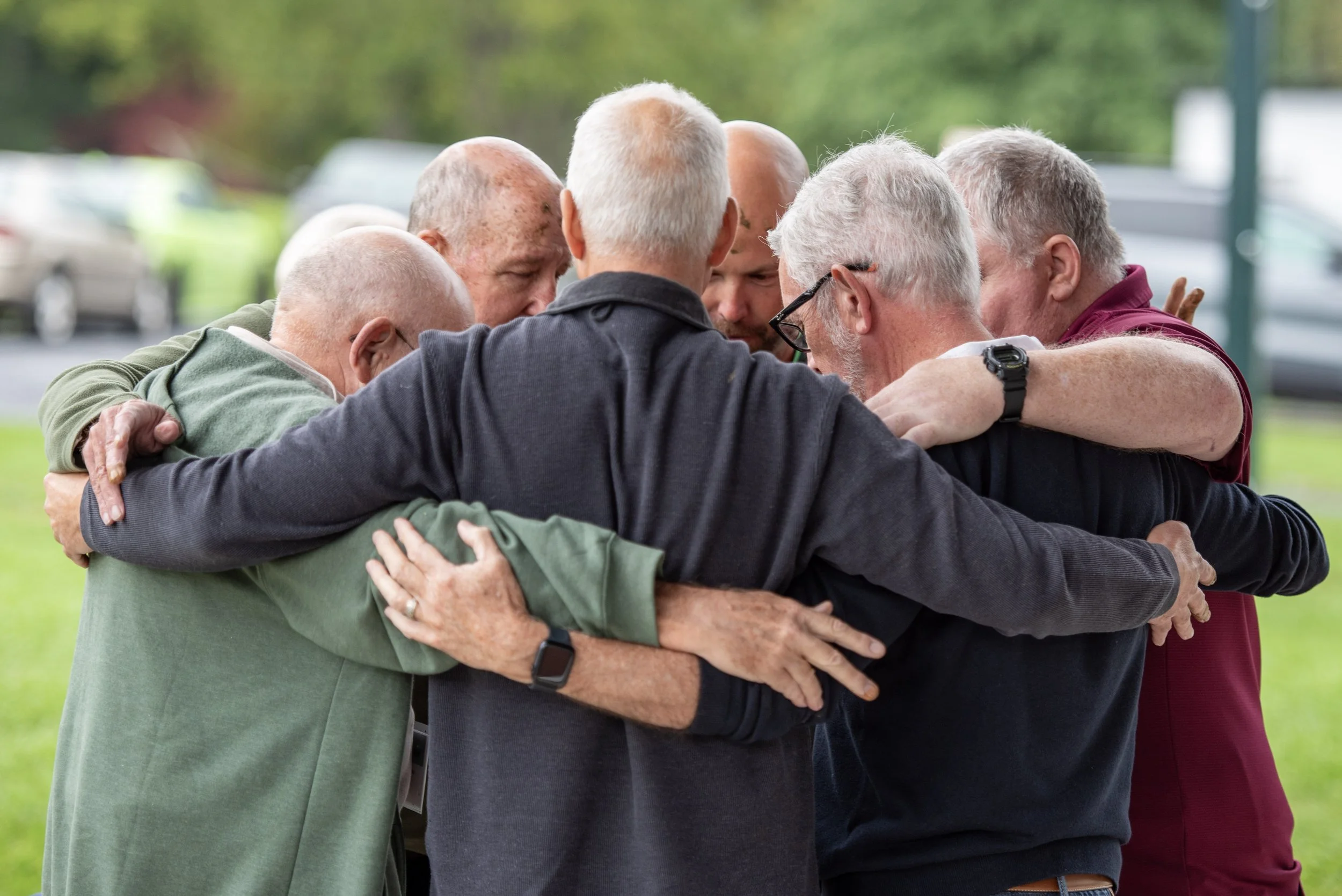Group of older men in a supportive huddle outdoors, embracing each other.