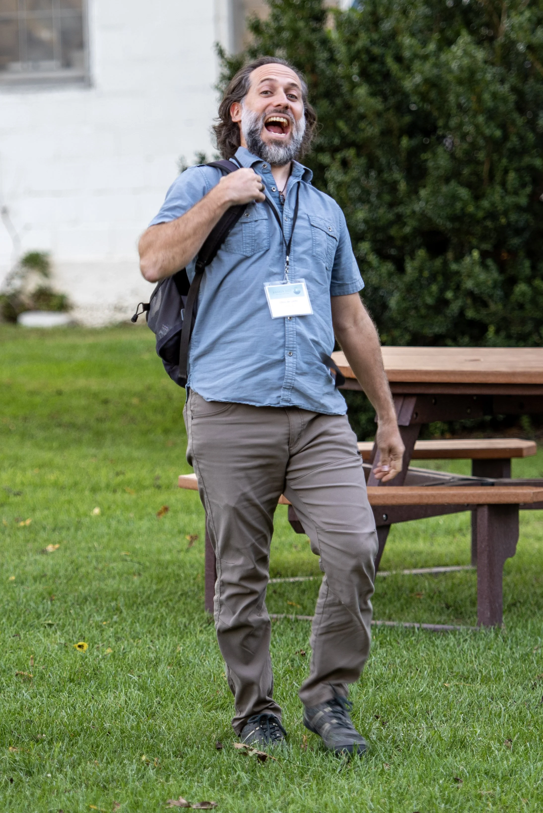 Man with a beard and backpack smiling and walking on grass near a picnic table.