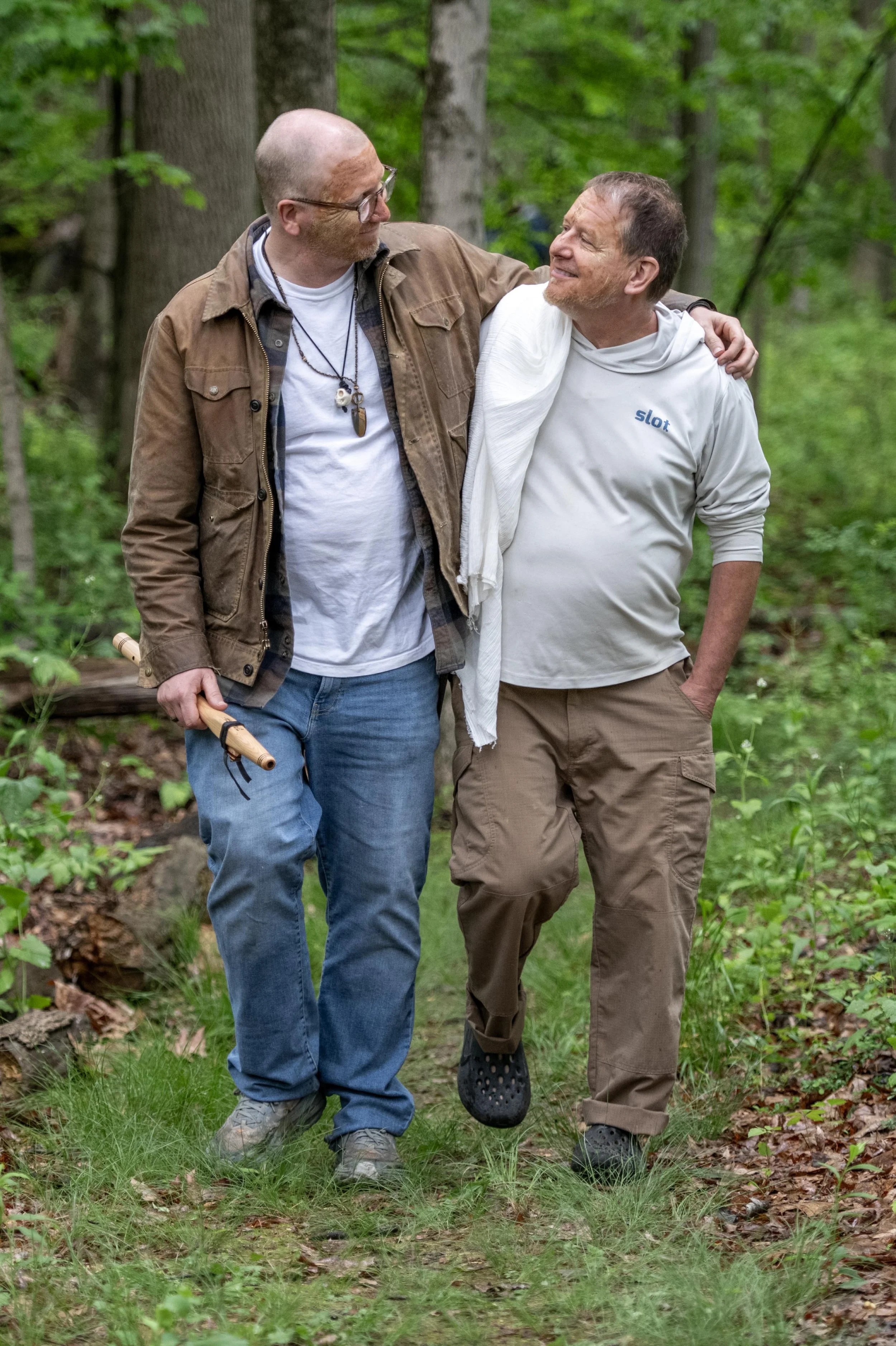 Two men walking in a forest, with one man helping the other walk by supporting his shoulder.