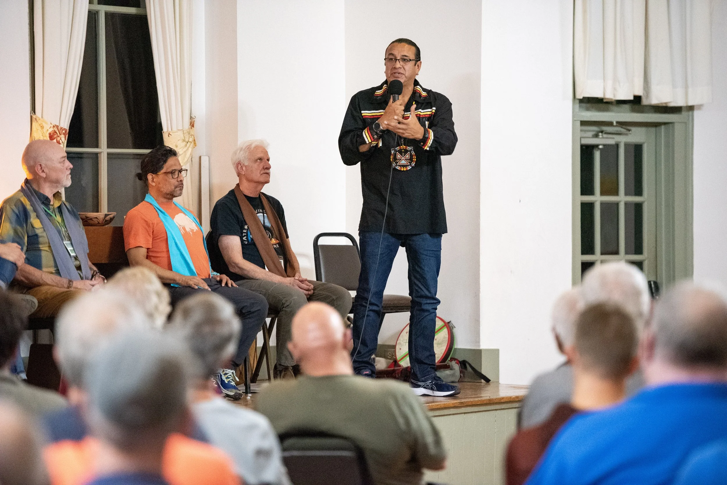 A person speaking in front of an audience at an indoor event while holding a microphone. Behind them, three seated individuals listen. The room features chairs, windows with curtains, and a large group of people facing the speaker.