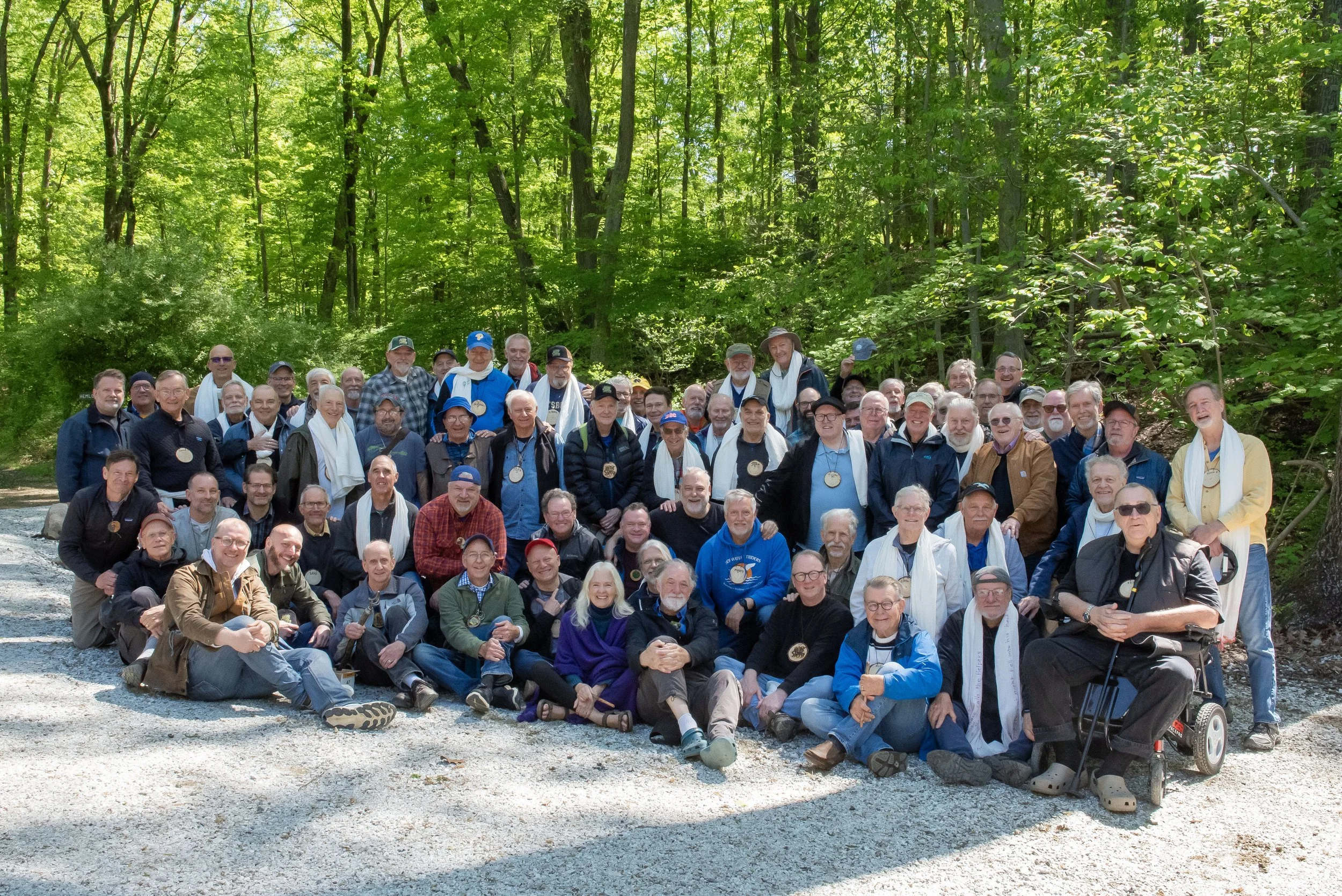 A large group of elderly people gathered outdoors in a forested area with lush green trees in the background, posing for a group photo on a gravel path.