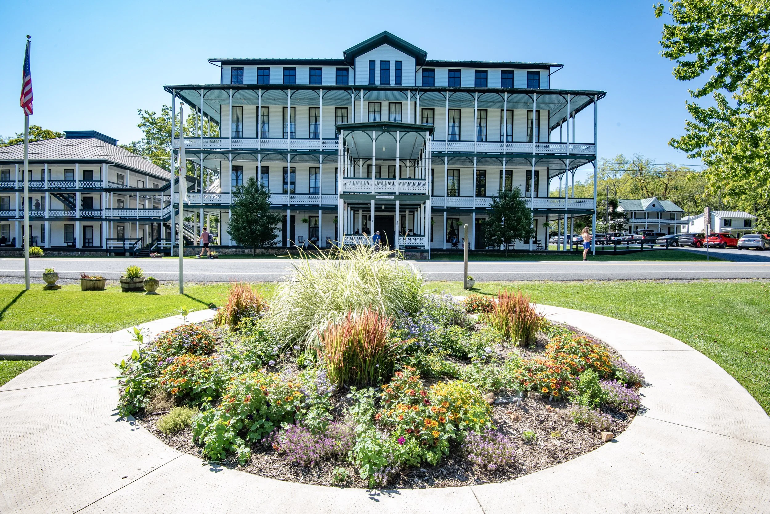 Historic hotel building with white facade and multiple balconies, surrounded by landscaped gardens with flowering plants, under a clear blue sky.
