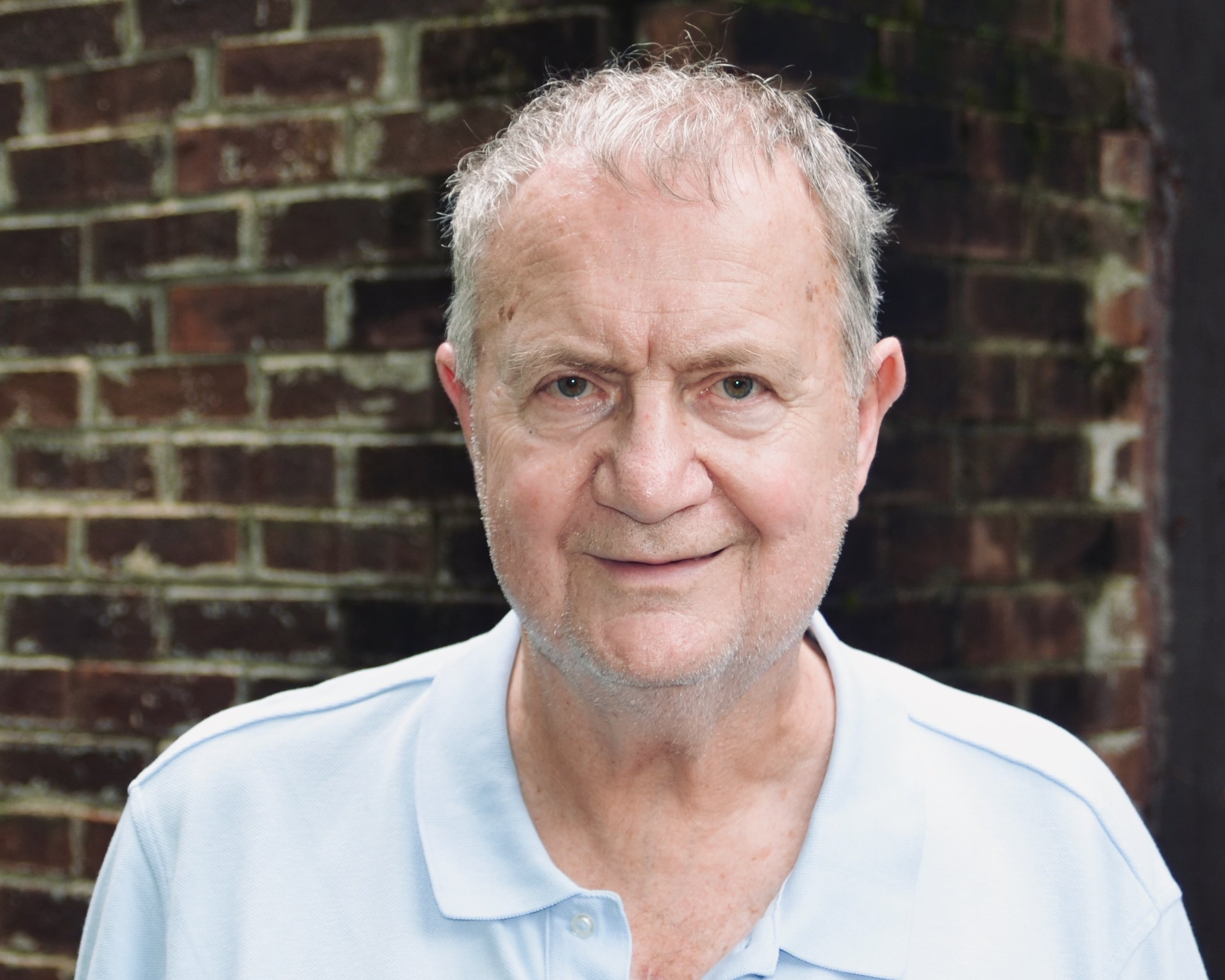 A smiling older man with short gray hair and a beard, wearing a red shirt, standing in front of a stone wall.