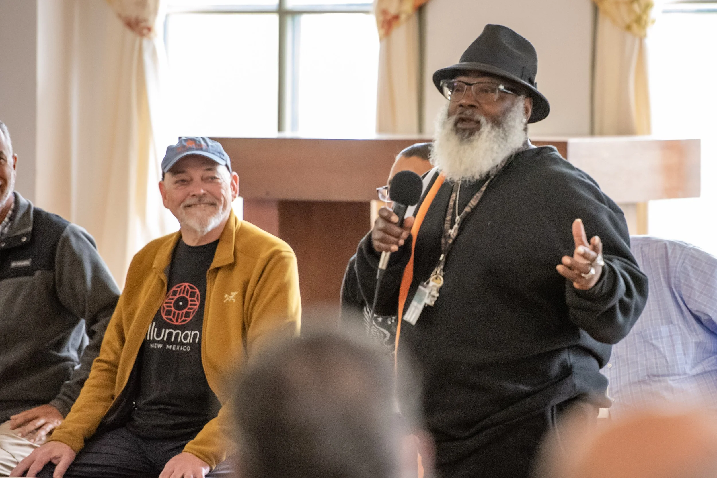 Two men speaking at an indoor event, one holding a microphone wearing a black hat, the other seated wearing a cap and jacket.