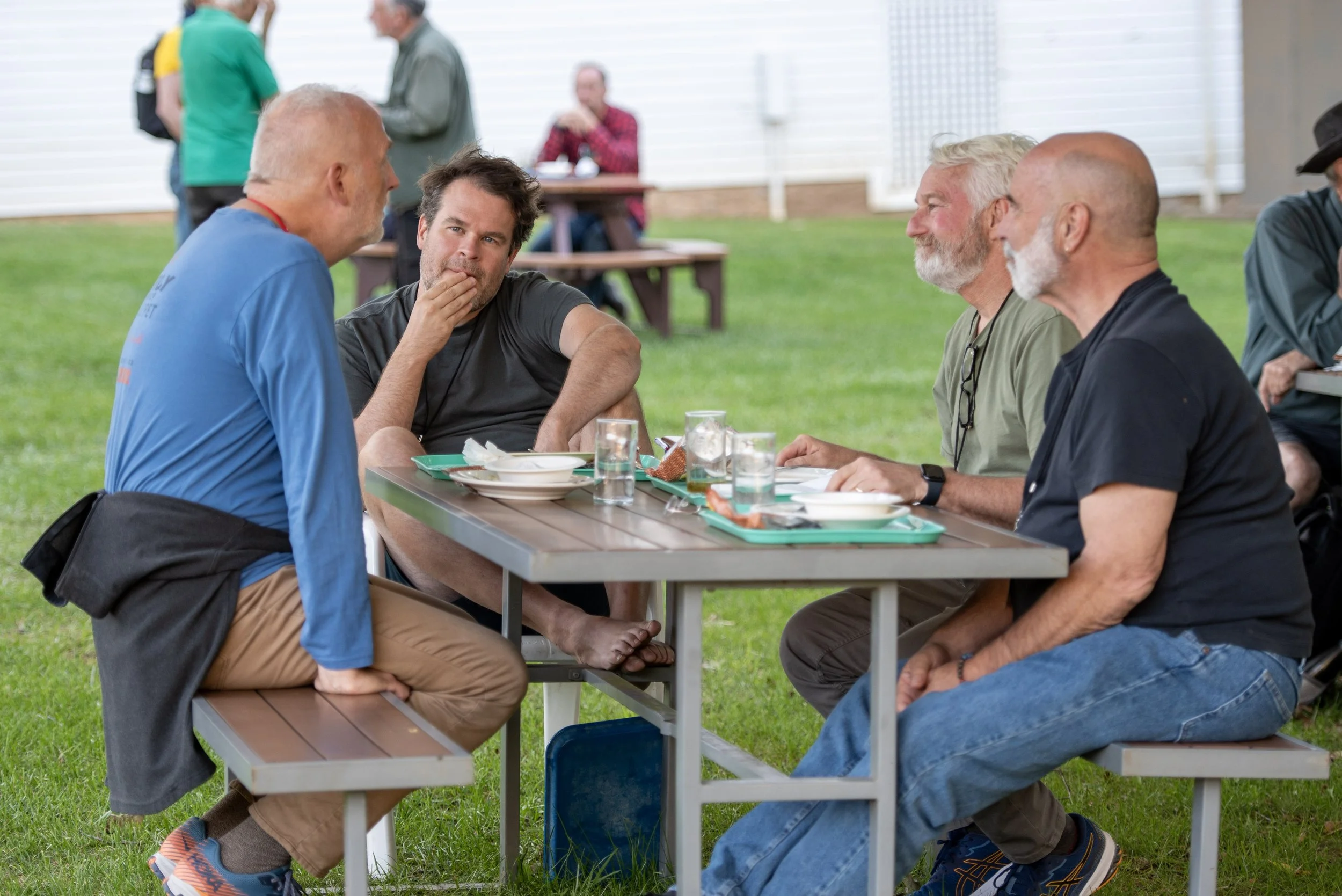 Four men sitting at a picnic table outdoors, having a conversation over food and drinks on a grassy area with people in the background.