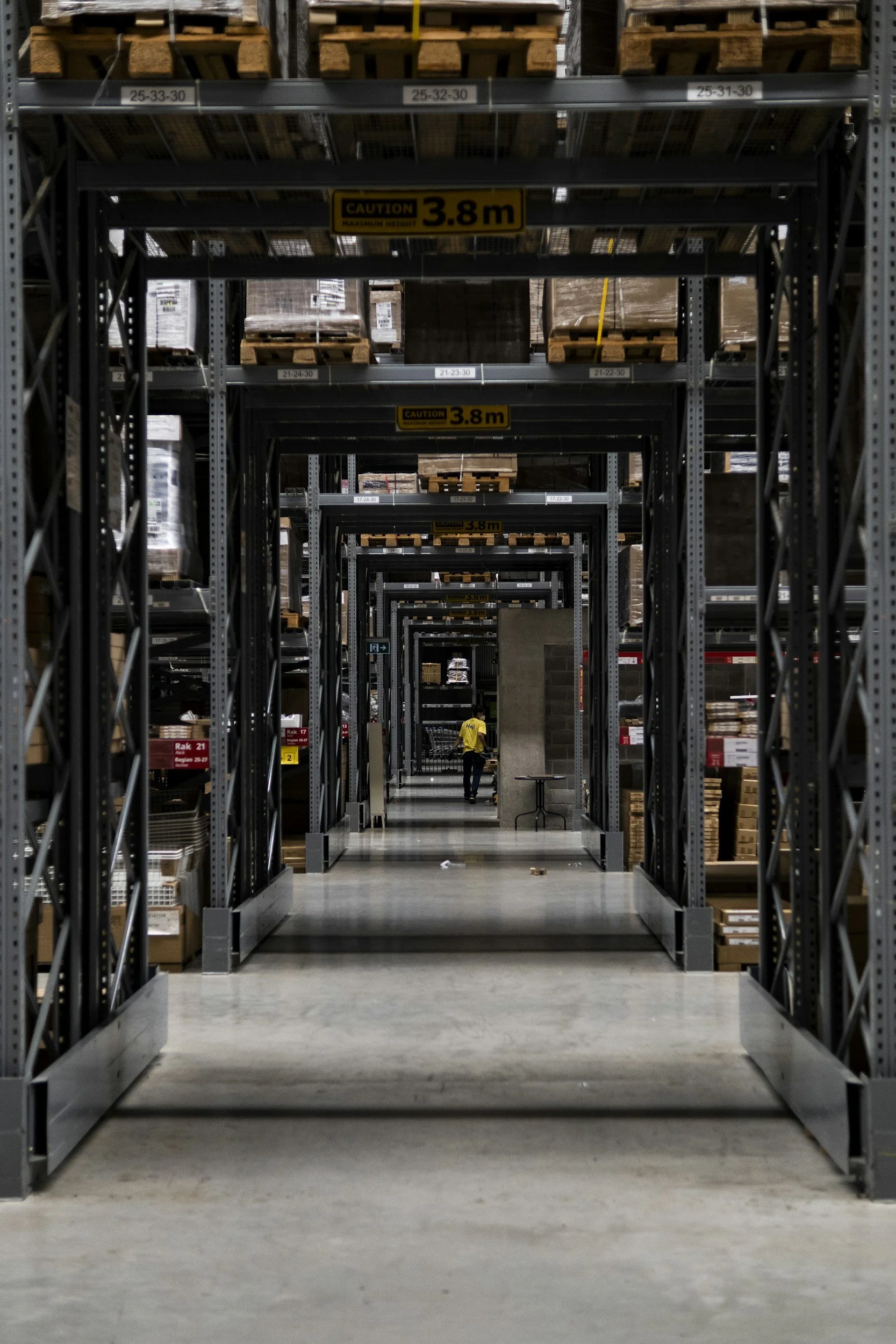 A warehouse aisle with metal shelving filled with boxes and pallets, marked with signs indicating a maximum height of 3.8 meters, and a person in a yellow shirt walking in the distance.