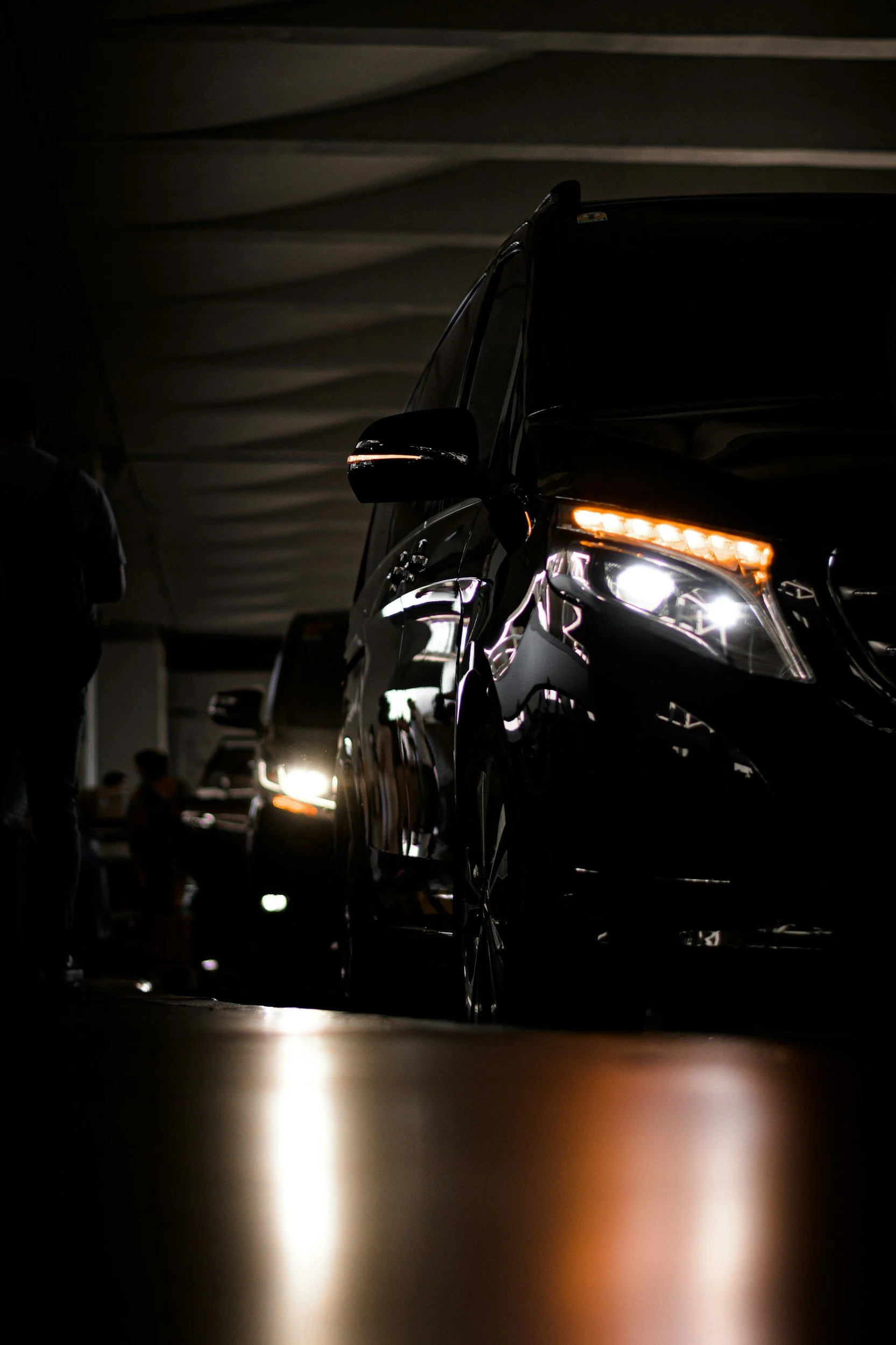 Black SUVs lined up indoors, with headlights on, in a dimly lit space. People are seen in the background.