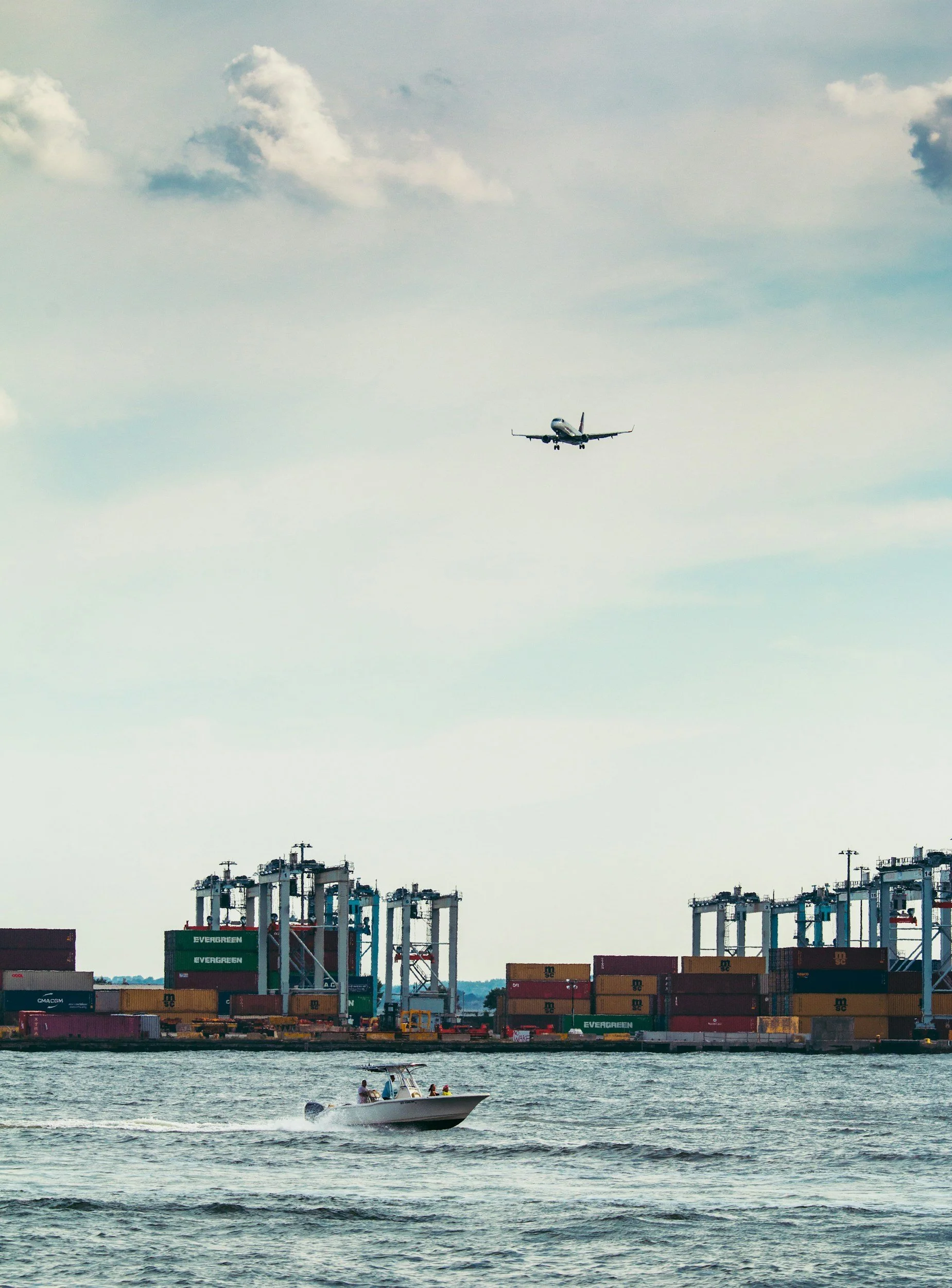 A plane flying over a harbor with container ships and a speedboat in the water.