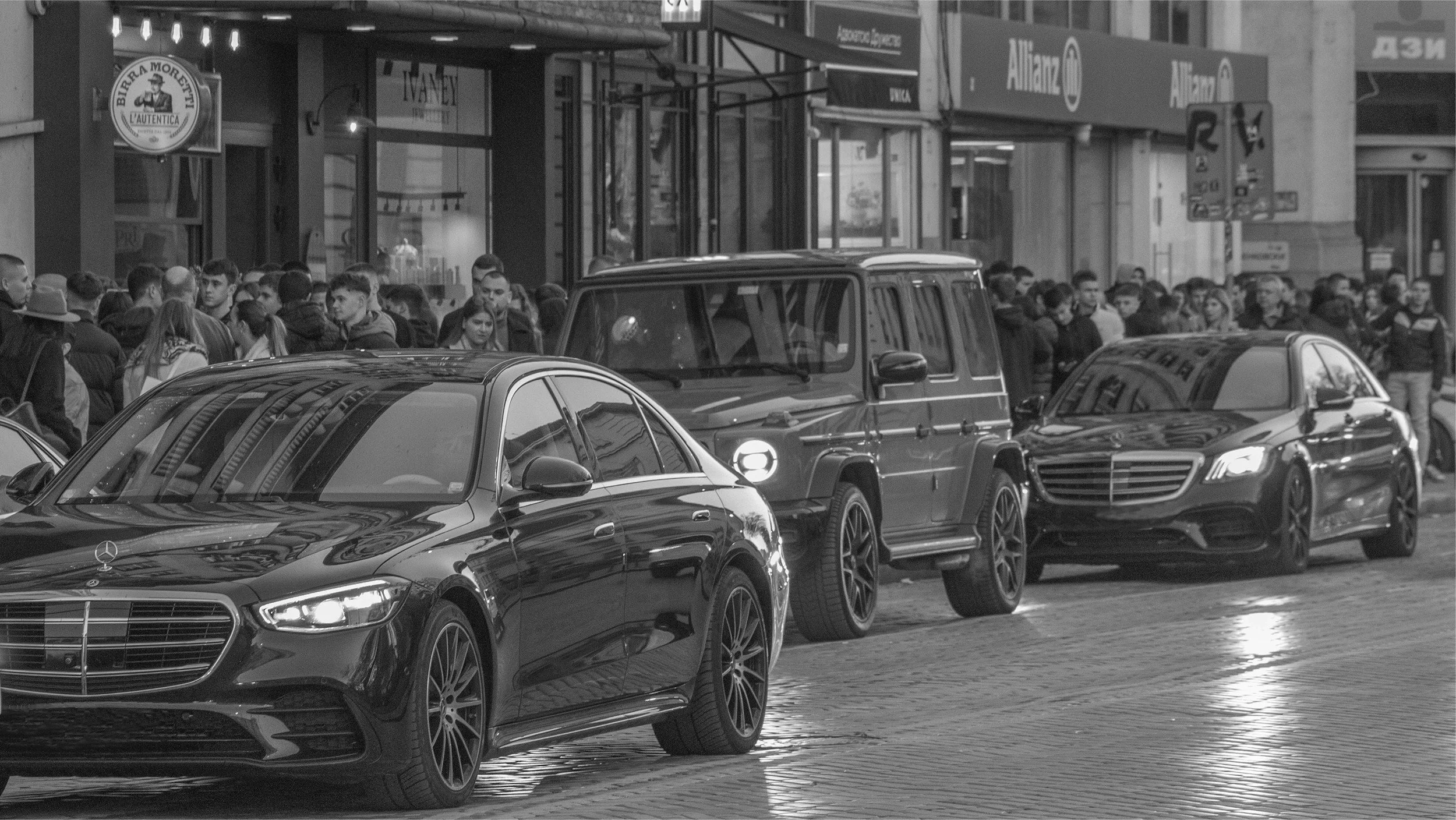 Black and white photo of a busy city street with three parked luxury cars: a Mercedes-Benz, a vintage Mercedes G-Class, and a modern Mercedes E-Class. In the background, there is a crowd of pedestrians walking on the sidewalk and storefronts with signs.