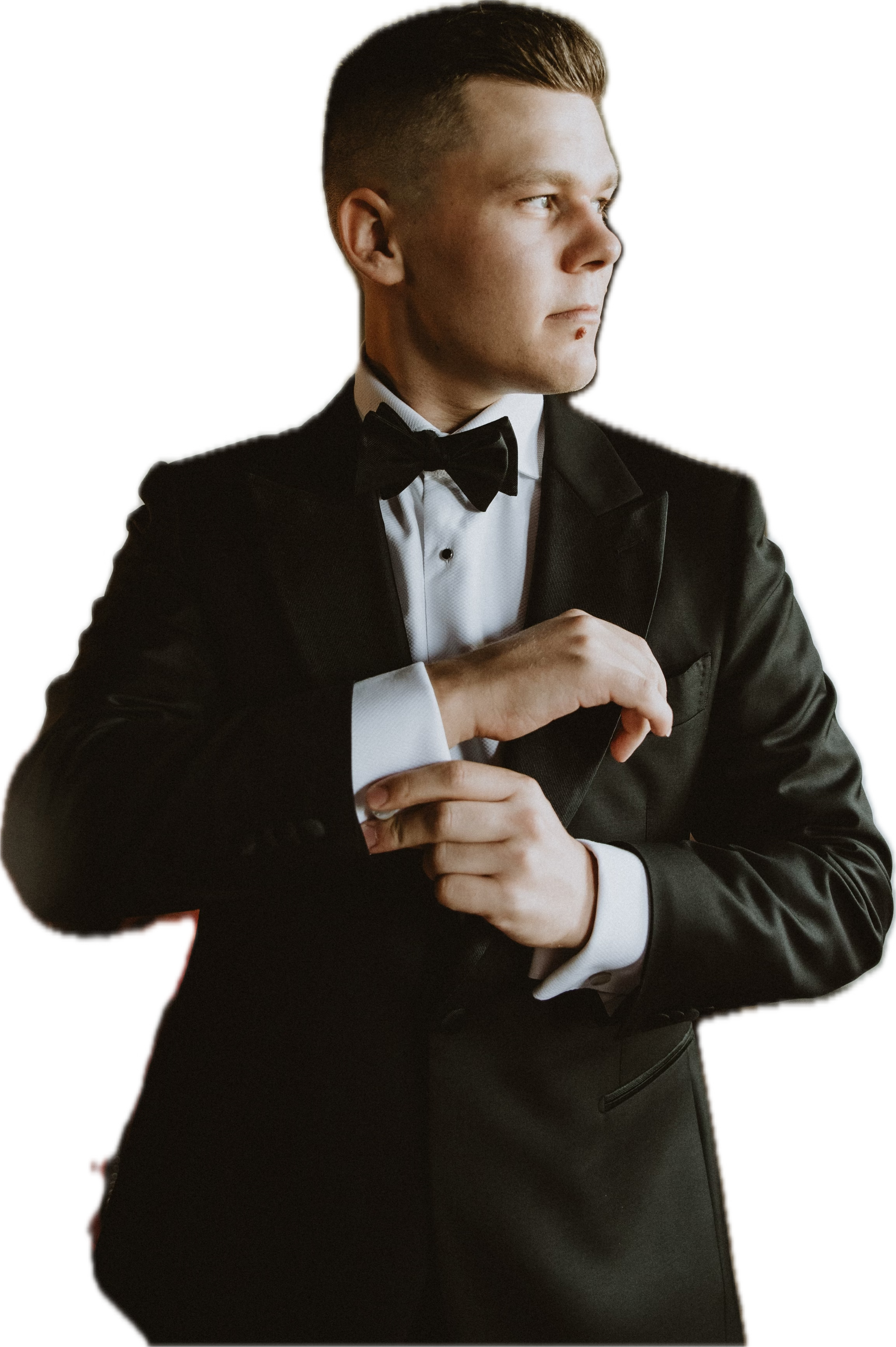 A young man in a black tuxedo with a bow tie is adjusting his sleeve, looking to the side.