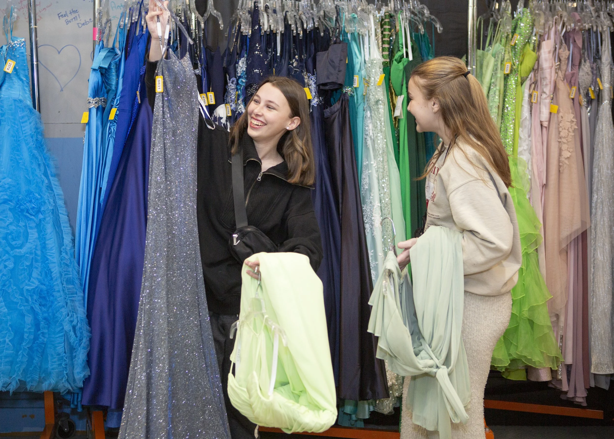 Two young women shopping for dresses in a store. They are surrounded by hanging dresses in various colors, including blue, silver, green, and pink.