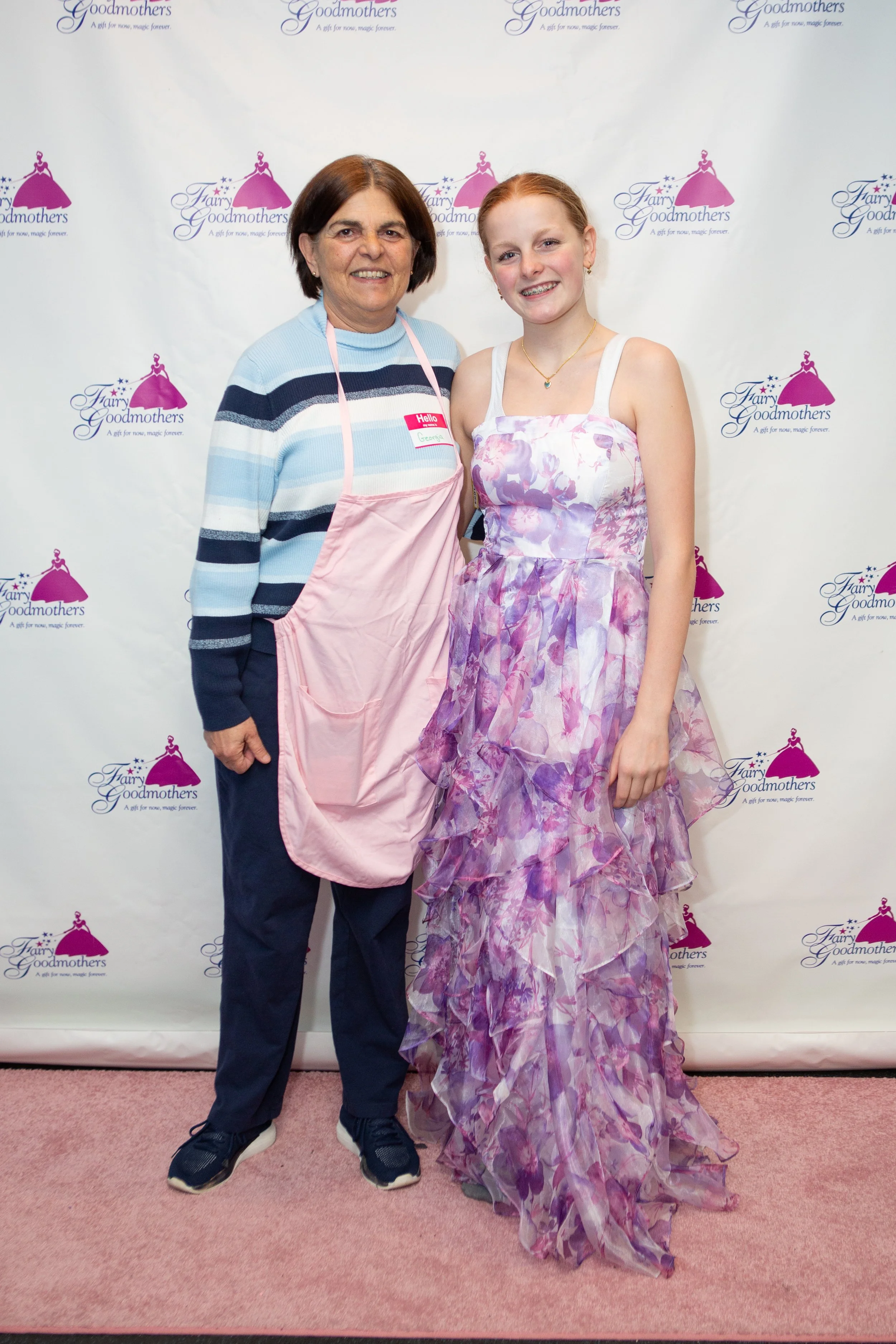 A woman and a girl standing in front of a backdrop with the Fairy Godmothers logo. The woman is wearing a striped sweater and an apron, and the girl is wearing a pink and purple floral dress with a necklace and braces, both smiling at the camera.