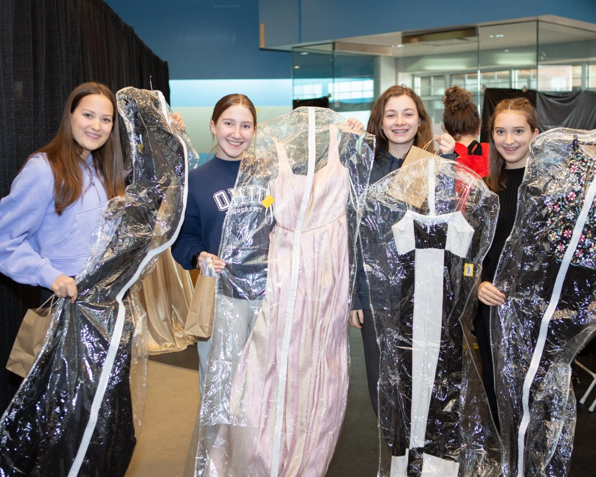 Group of young girls smiling and holding clear plastic garment covers with dresses inside, standing indoors in a bright, modern space.