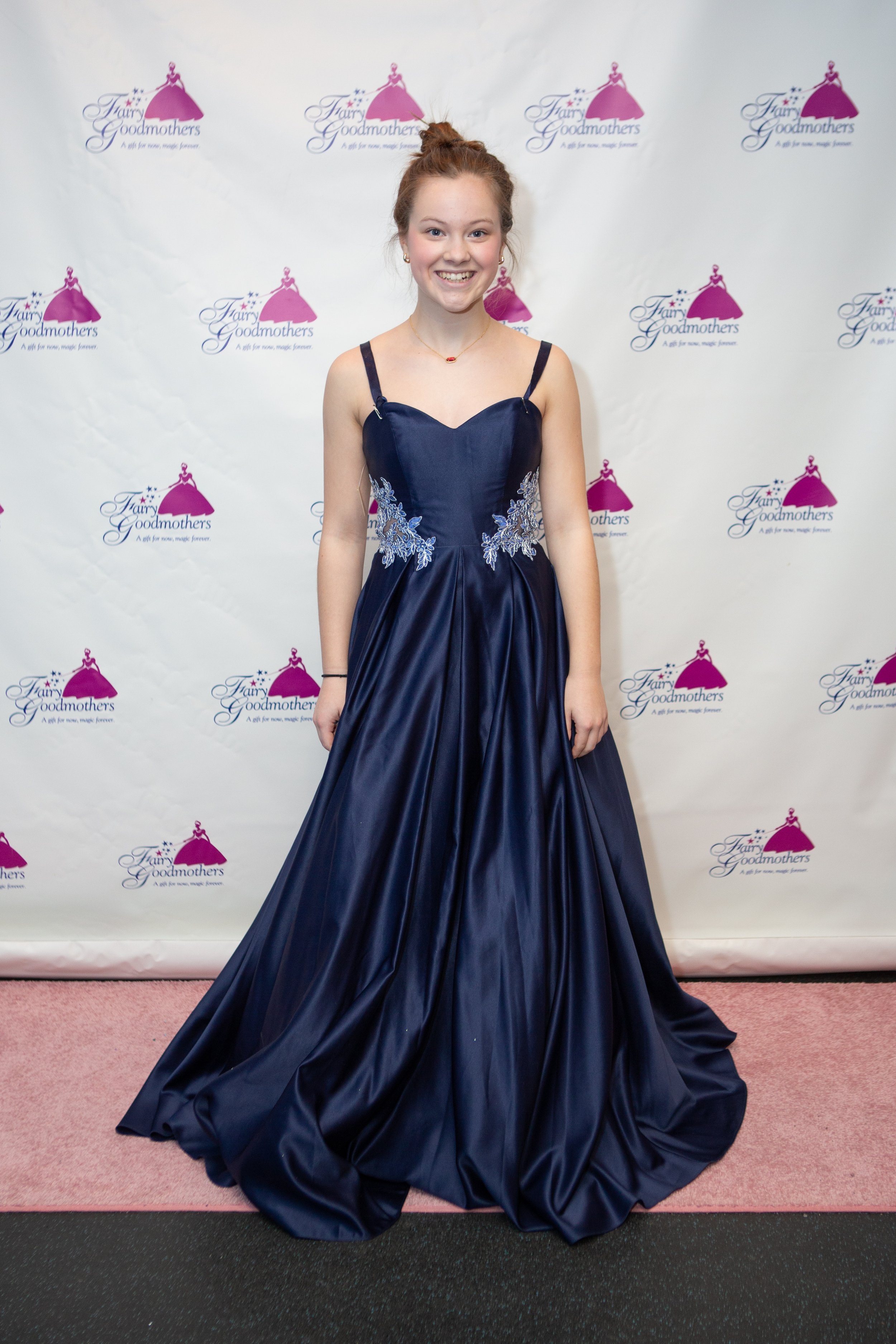 Young woman in a navy blue formal gown standing in front of a backdrop with 'Fairy Godmothers' logo, smiling at the camera.