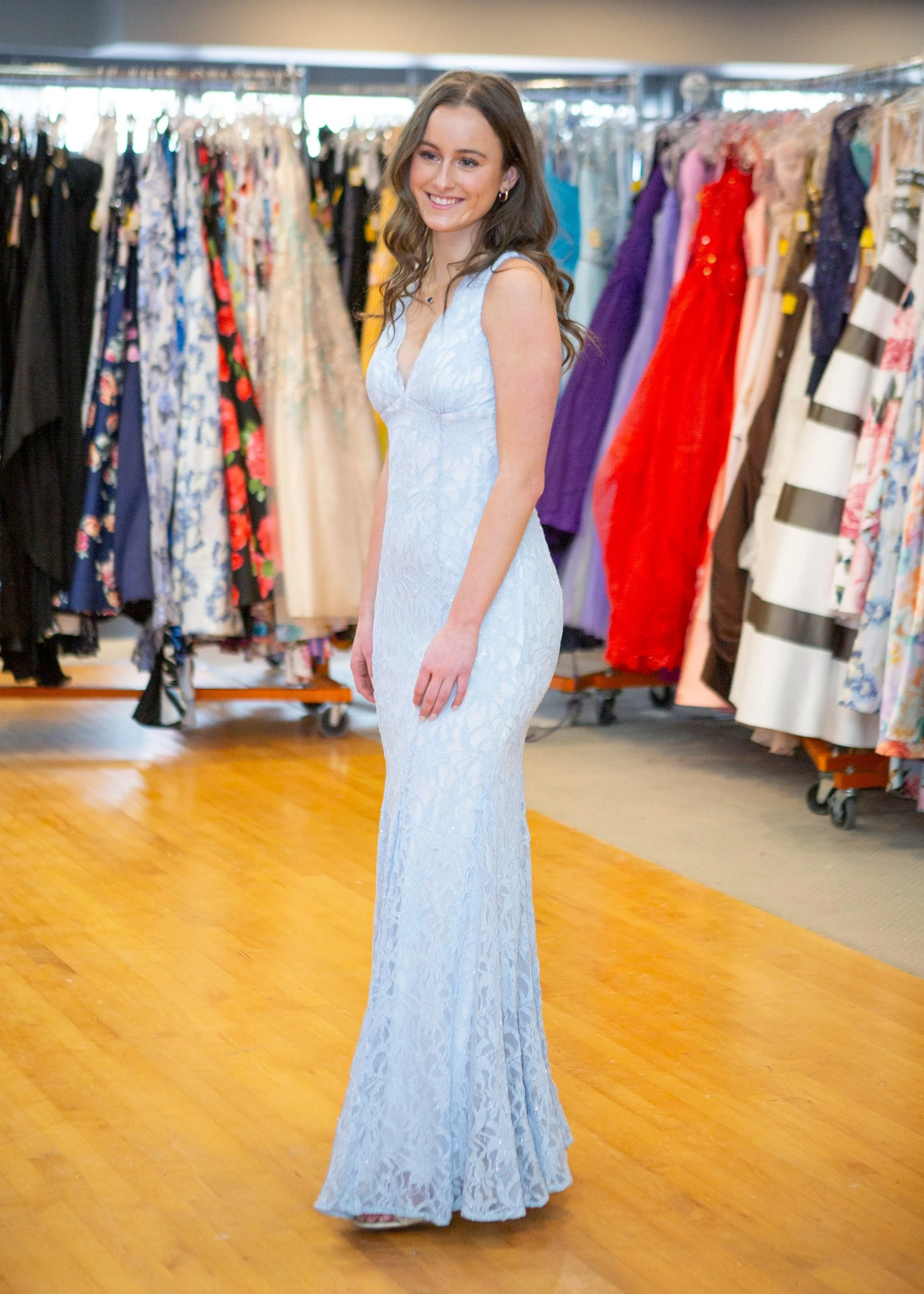 A woman in a light blue lace dress trying on a dress inside a clothing store, surrounded by racks of colorful dresses.