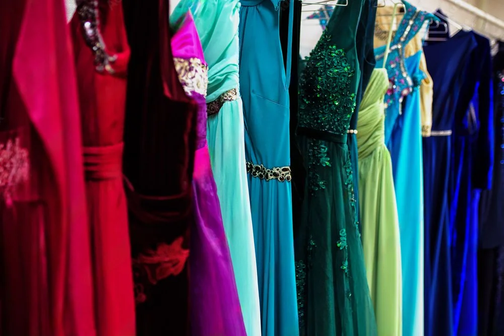 A row of colorful evening gowns hanging on a clothing rack, featuring various shades of red, purple, blue, green, and yellow, with some dresses embellished with sequins or embroidery.